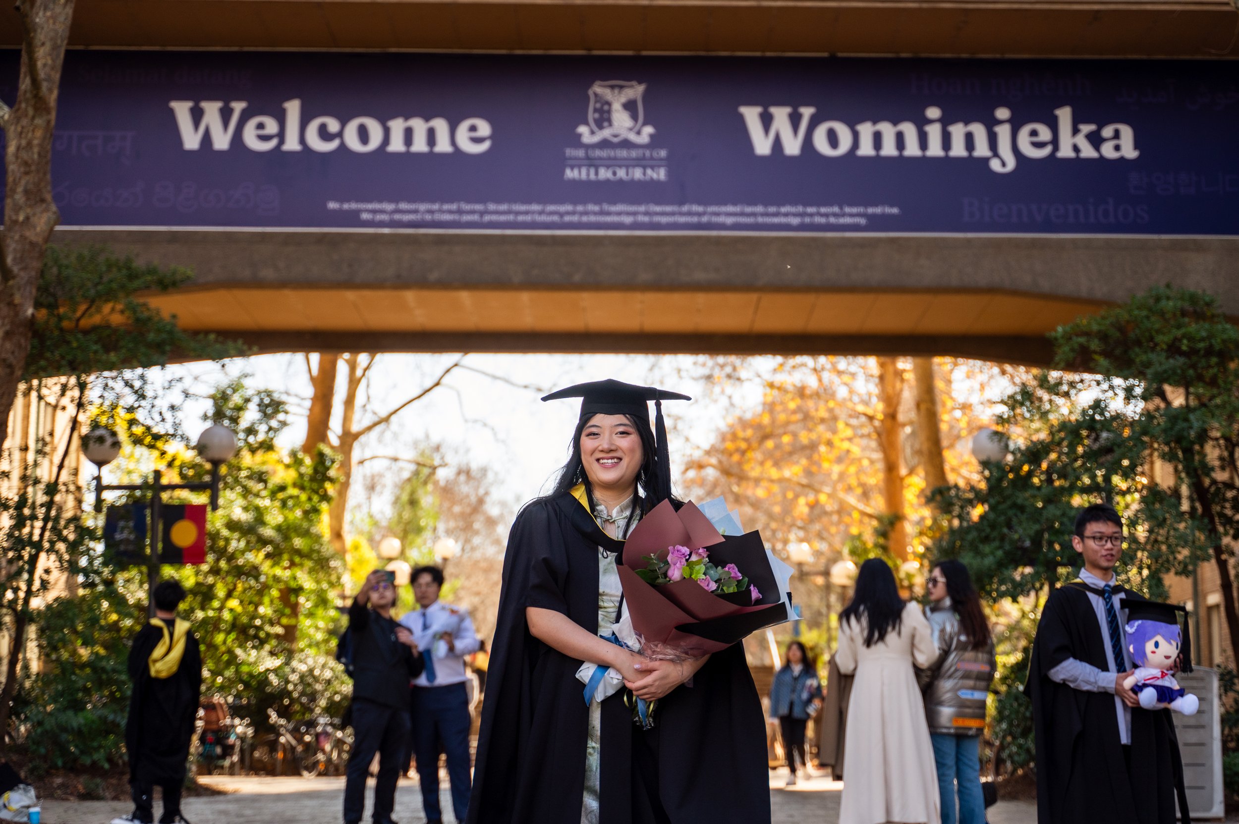 A young woman in graduation attire holding a bouquet of flowers, smiling at a university graduation ceremony outdoors with several other graduates and attendees in the background.