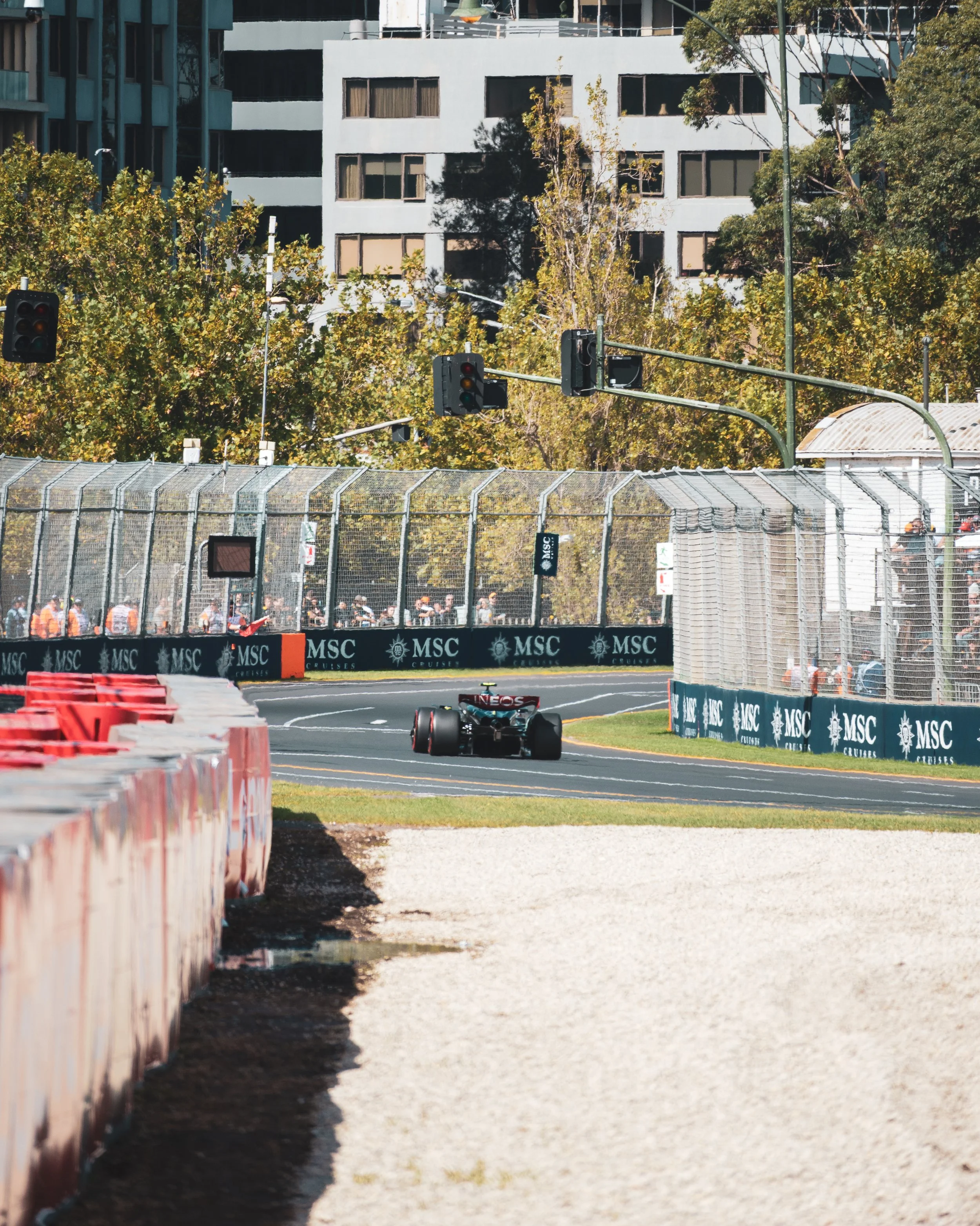 A black Formula 1 race car on a street circuit, with safety barriers, fencing, and traffic lights, surrounded by trees and tall buildings.