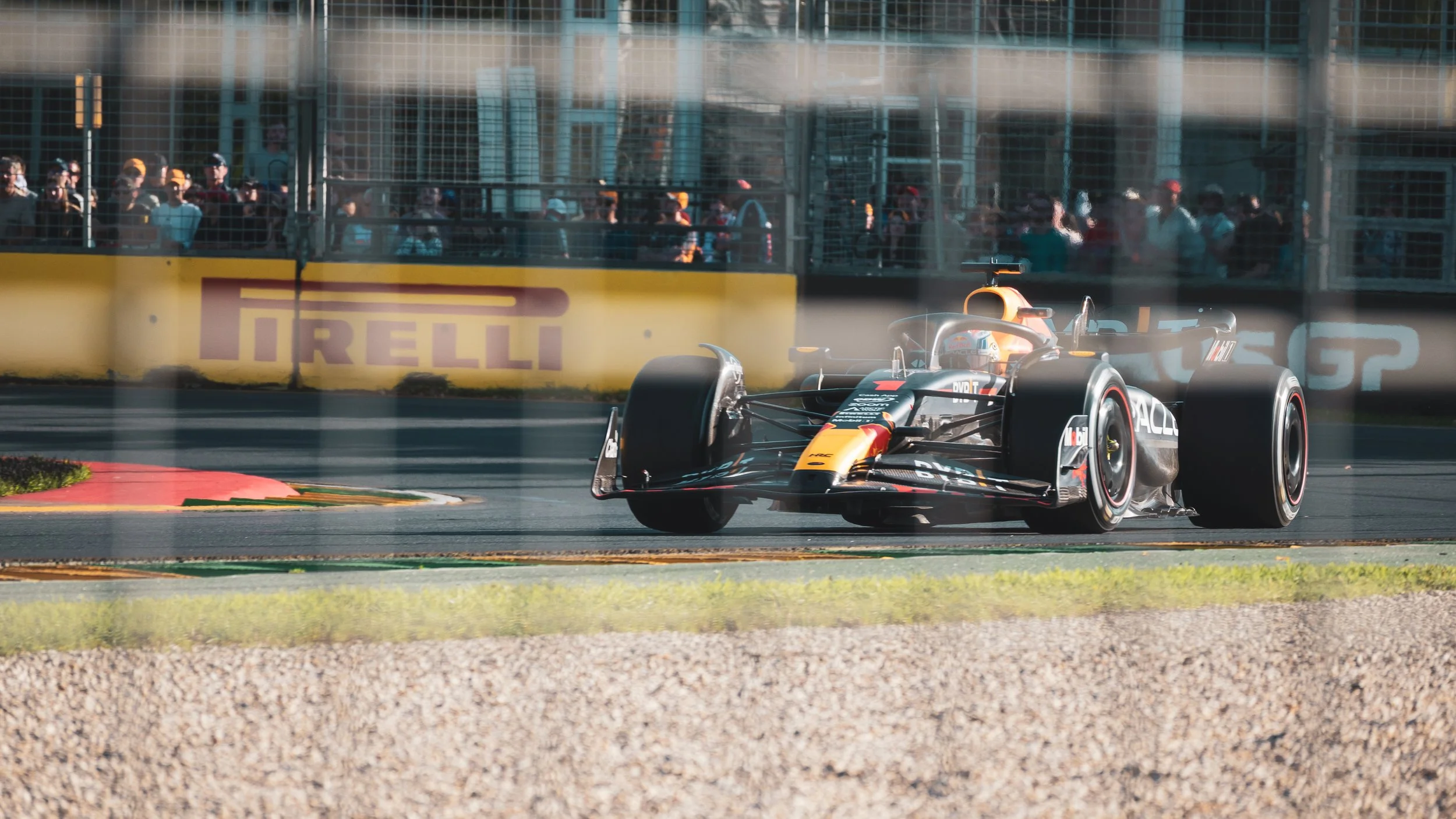 A race car speeding around a corner on a racetrack with spectators watching behind a glass barrier, Pirelli advertising banner in the background.