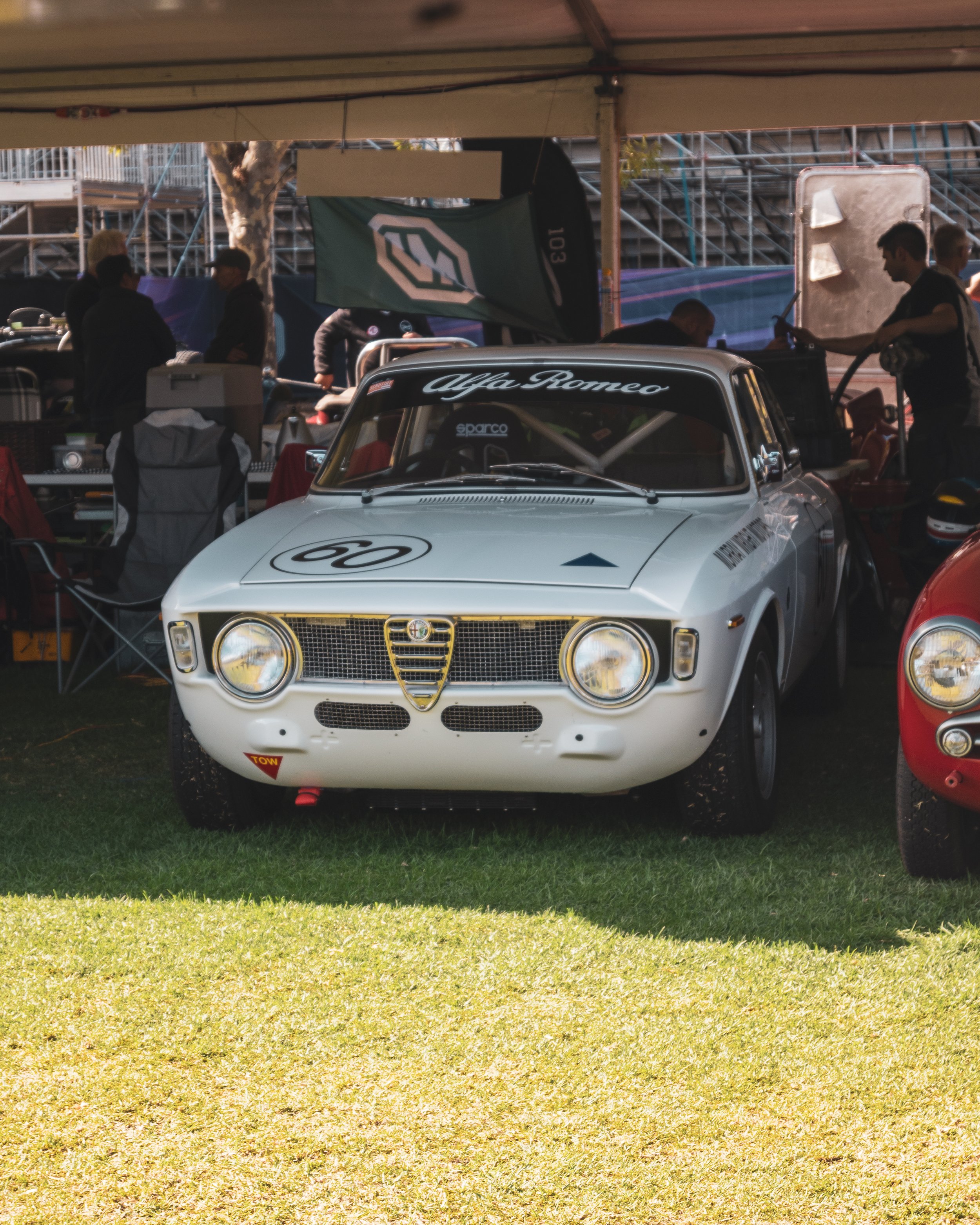White vintage Alfa Romeo race car with the number 60 on the hood, parked under a tent at a racing event, with people working and spectating in the background.
