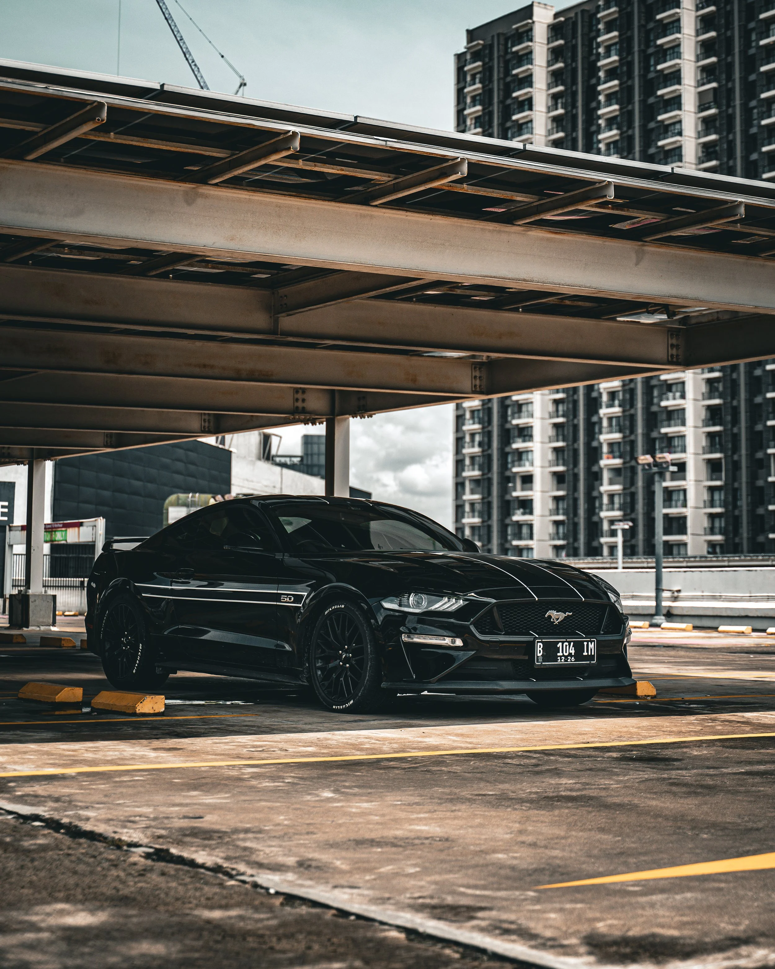 A black Ford Mustang car parked in an outdoor parking lot beneath a concrete overpass with high-rise buildings in the background.