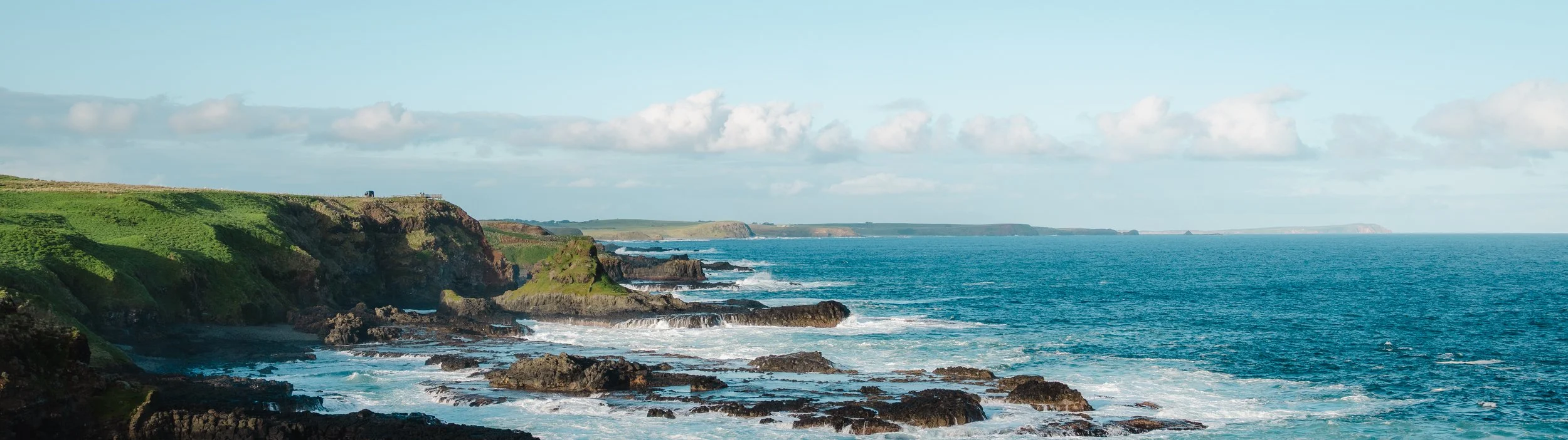Scenic view of a rugged coastline with green cliffs, rocks, and ocean waves under a partly cloudy sky.