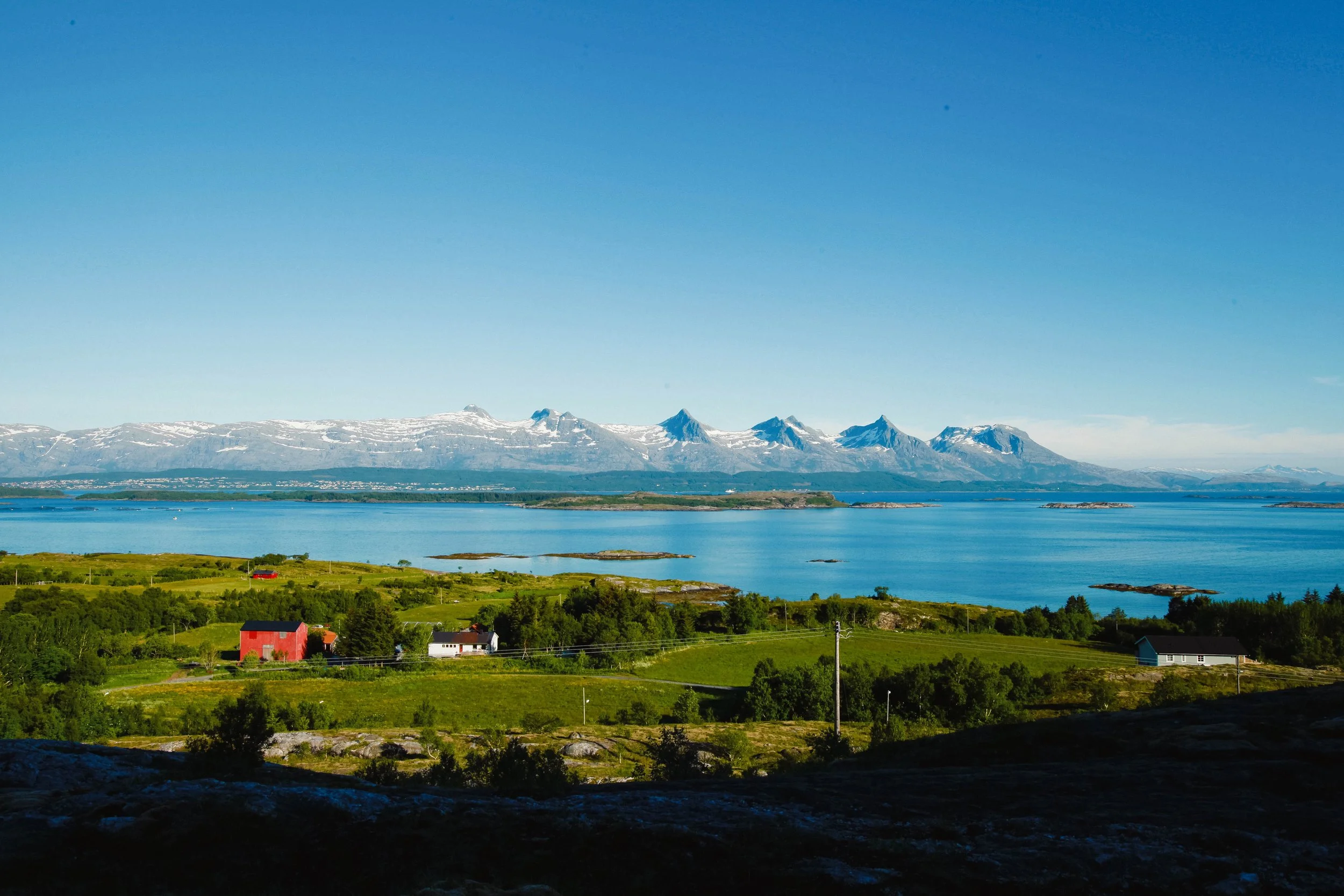 Fjellrekka De syv søstre med snøkledde topper, sett over havet fra kysten utenfor. Grønne jorder, hus og holmer i forgrunnen under klar blå himmel. Bildet er tatt fra Herøy.
