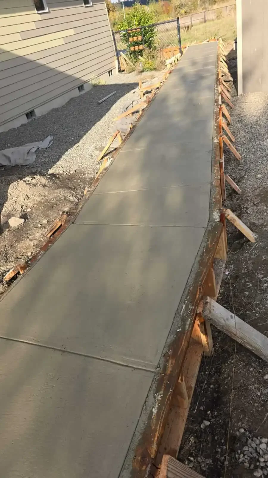 Concrete sidewalk under construction, with wooden forms supporting the poured concrete, adjacent to a beige house with horizontal siding.