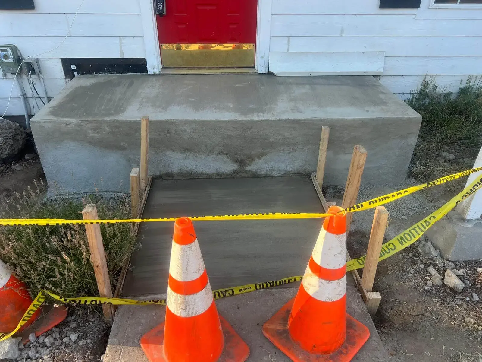 Concrete porch with new poured step, surrounded by construction cones and caution tape, in front of a house with a red door.