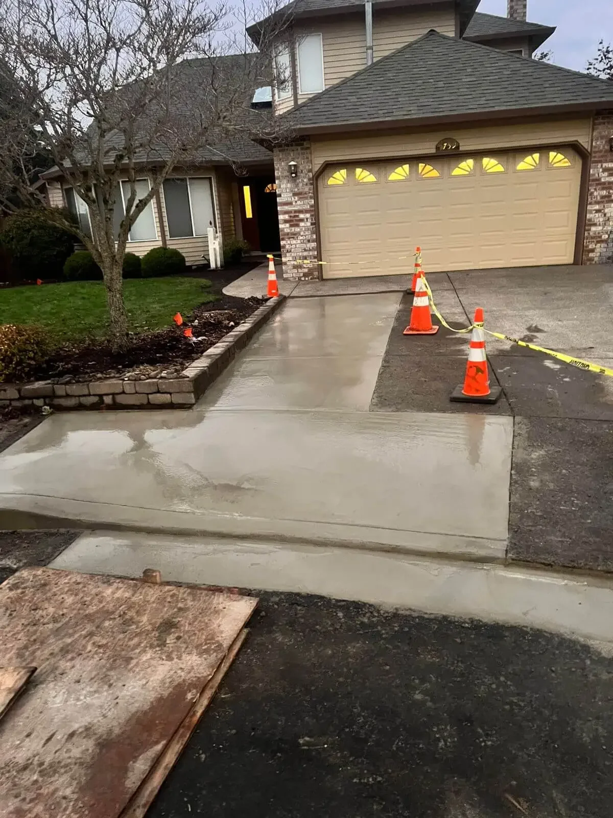 New concrete driveway being poured and cured in front of a house, with orange construction cones and caution tape around work area.