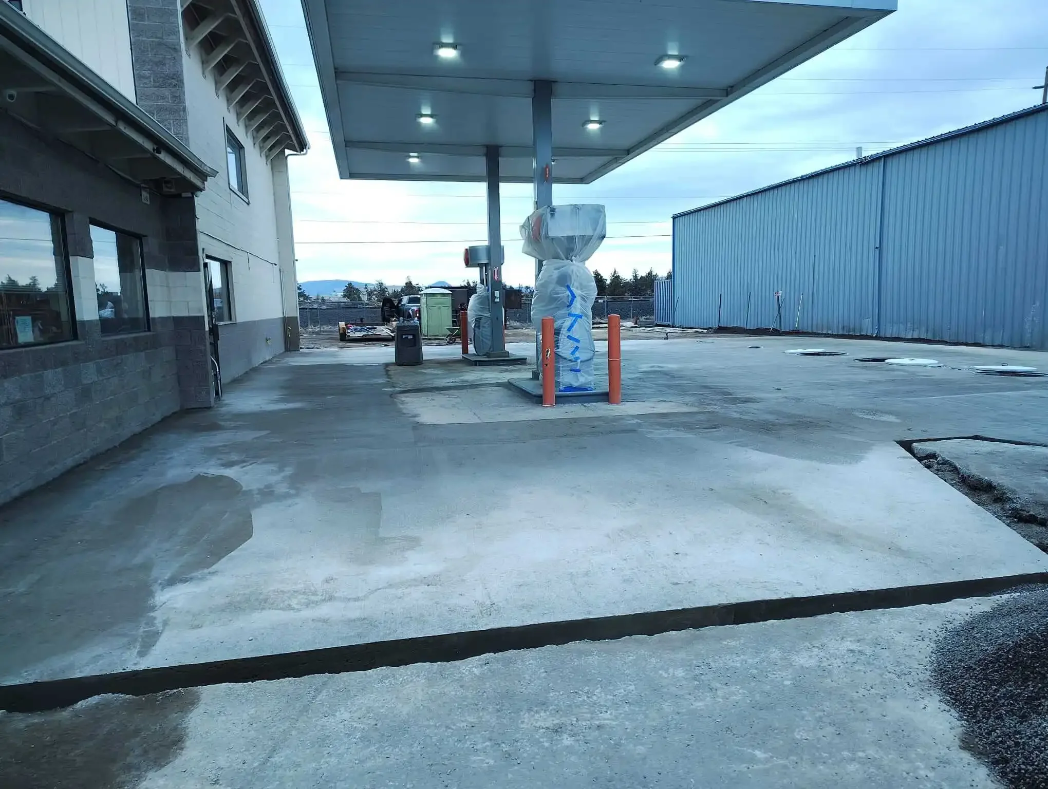 Empty parking lot outside a building with a gas station canopy, covered fuel pumps, and construction materials in the background.