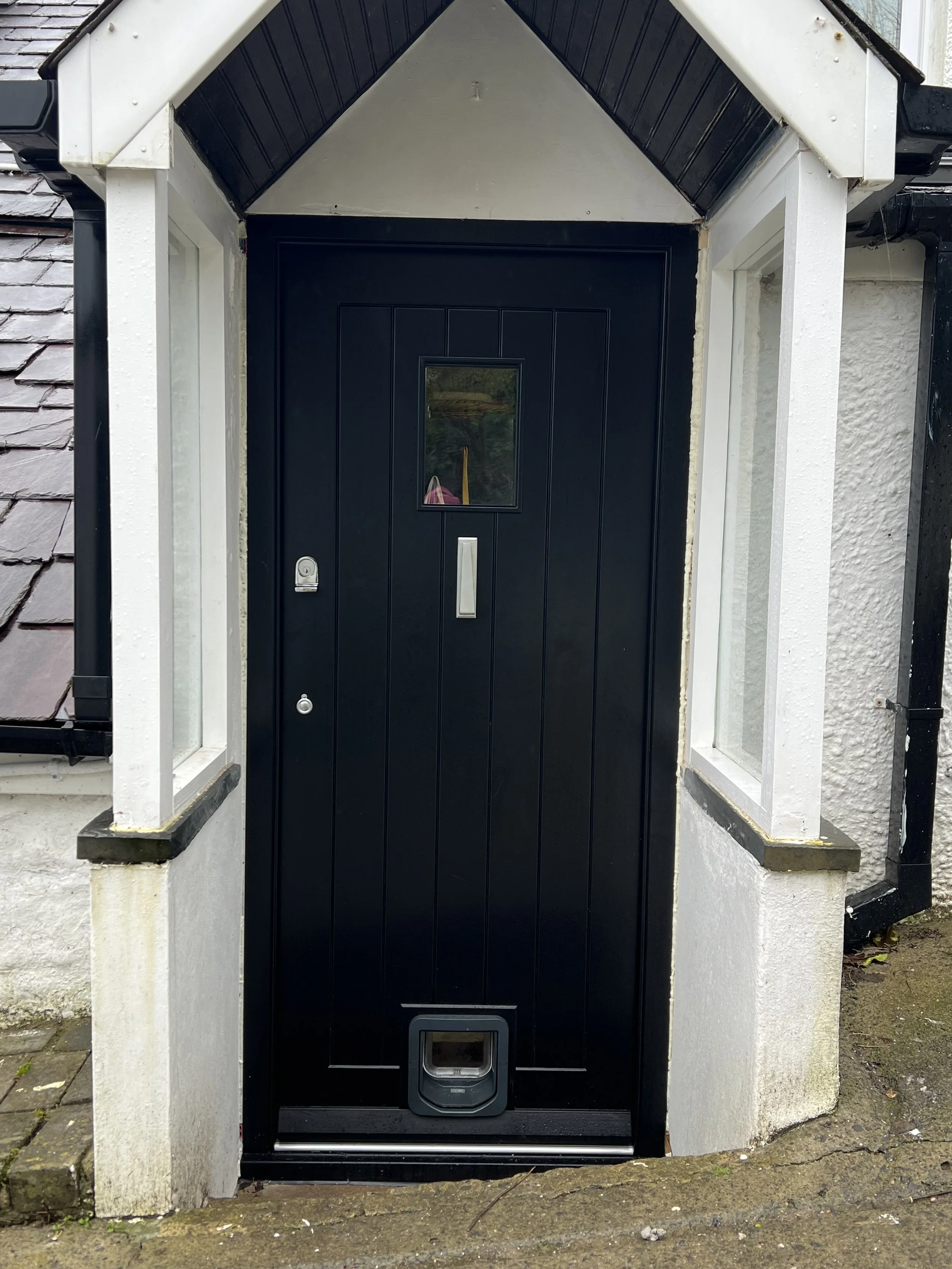 Black front door with a small window and cat flap at the bottom, flanked by white walls with windows on each side, leading to a porch with stone and brick paving.