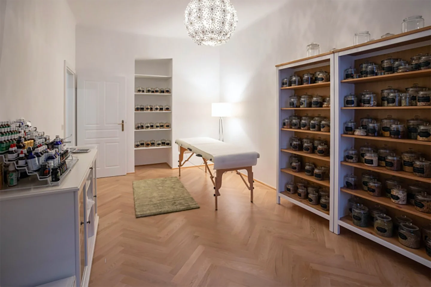 Ein Raum mit weißen Wänden, Holzfußboden, mehreren Regalen voller Gläser und einer Liege in der Mitte, verwendet wahrscheinlich für Massage oder Therapien.