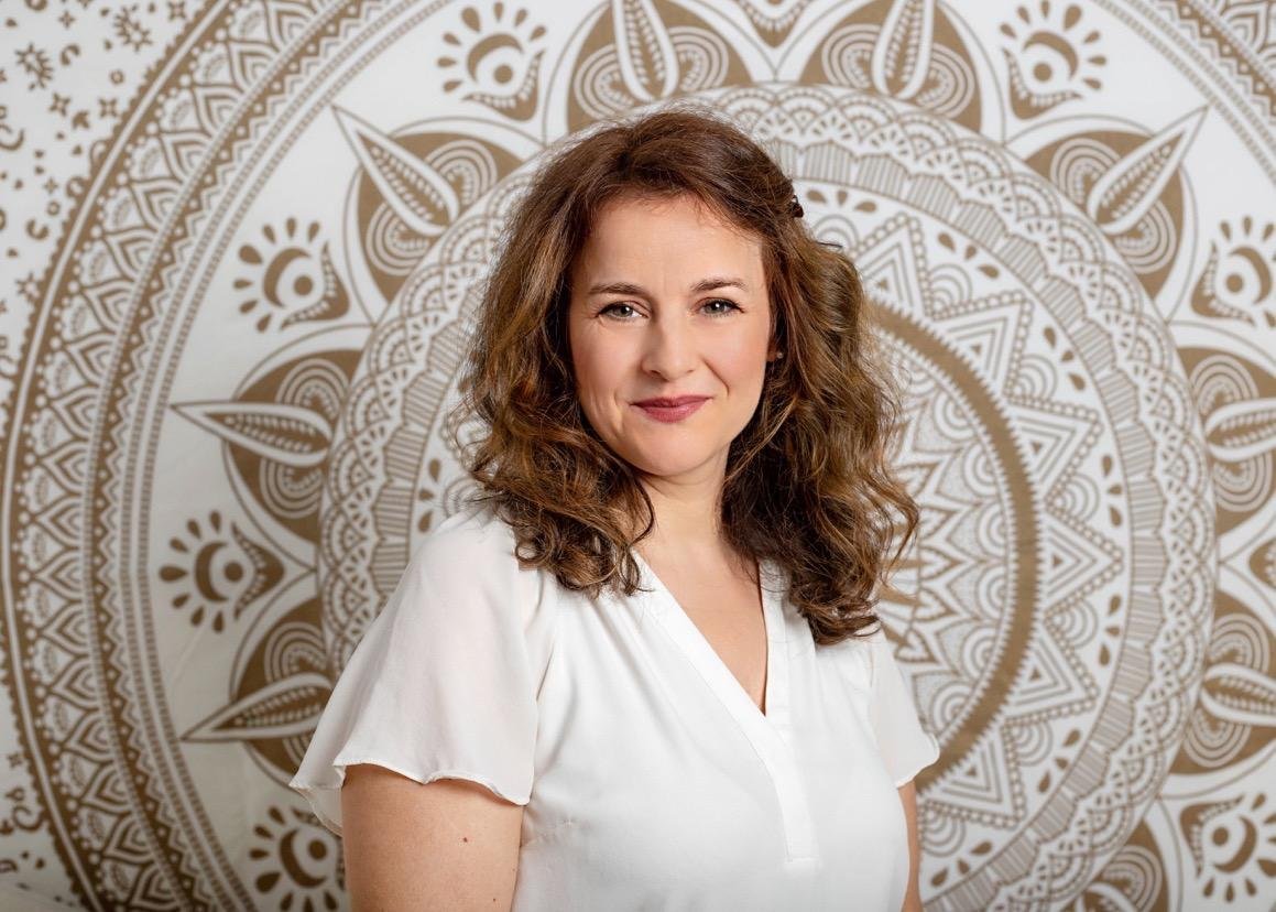 Eine Frau mit braunen, lockigen Haaren in einem weißen Oberteil vor einem gold-weiß Muster-Hintergrund.