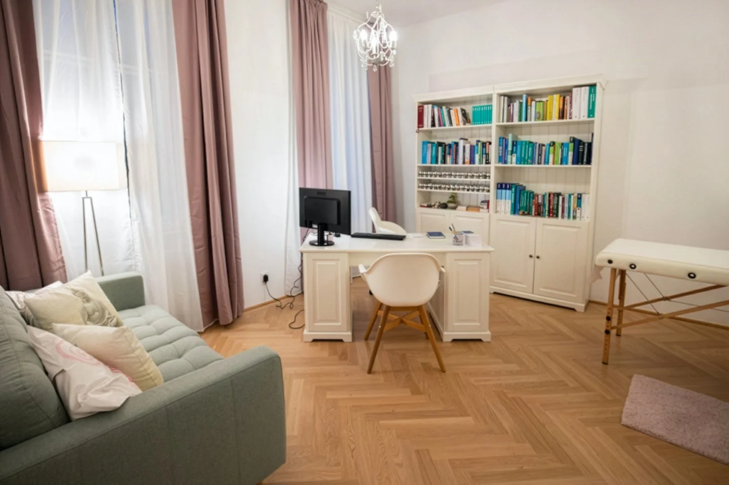 Ein helles, gut organisiertes Büro mit einem weißen Schreibtisch, einem Computer und einem Stuhl. Im Hintergrund eine weiße Bücherei und ein Kristallkronleuchter. Auf der linken Seite ein graues Sofa mit Kissen, an der Wand ein Lampenständer. Rechte Seite eine Behandlungsliege, auf dem Boden ein kleiner Teppich.