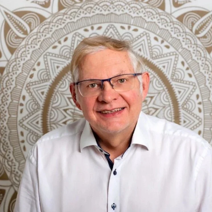 Ein Mann mit Brille und weißem Hemd vor einem Ornament-Hintergrund.