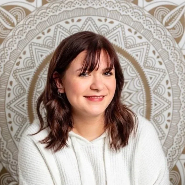 Junge Frau mit braunen, gewellten Haaren und weißen Pullover vor einer mandalaartigen Wanddekoration.