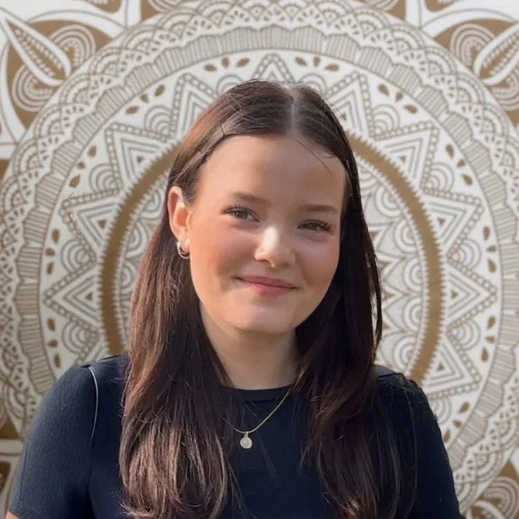 Junge Frau mit longem braunem Haar, lächelnd, steht vor einer Rose und weißen Mandala-ähnlichen Wanddekoration.