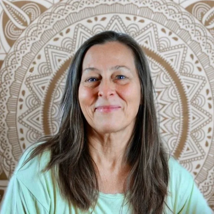 Eine lächelnde Frau mit schulterlangem braunem Haar vor einem Mandala-Hintergrund, trägt ein hellgrünes Oberteil.