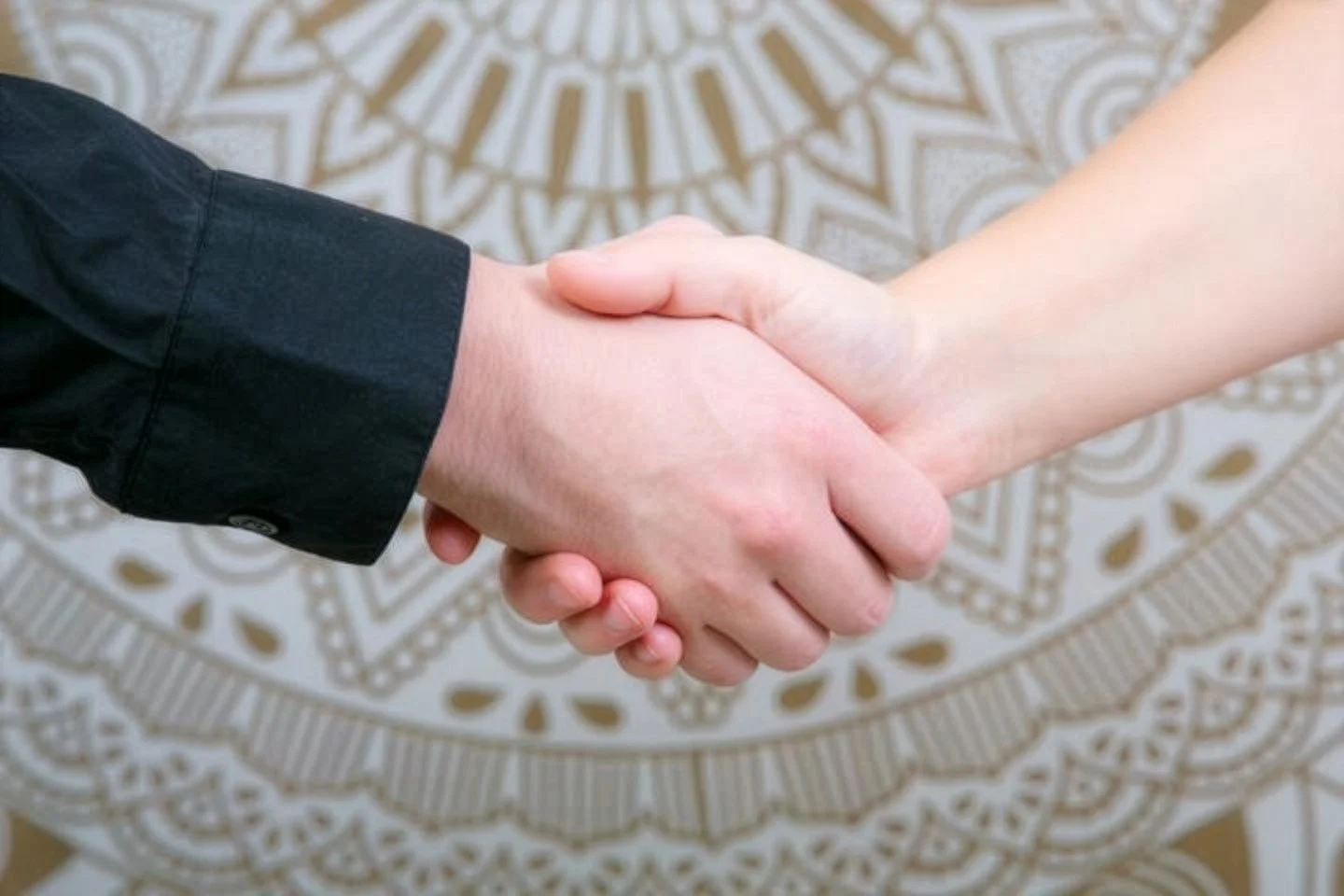 Nahaufnahme eines Händeschüttelns zwischen einer Person in einem schwarzen Hemd und einer in einem kurzen Ärmel shirt, vor einem dekorativen, goldfarbenen Muster im Hintergrund.