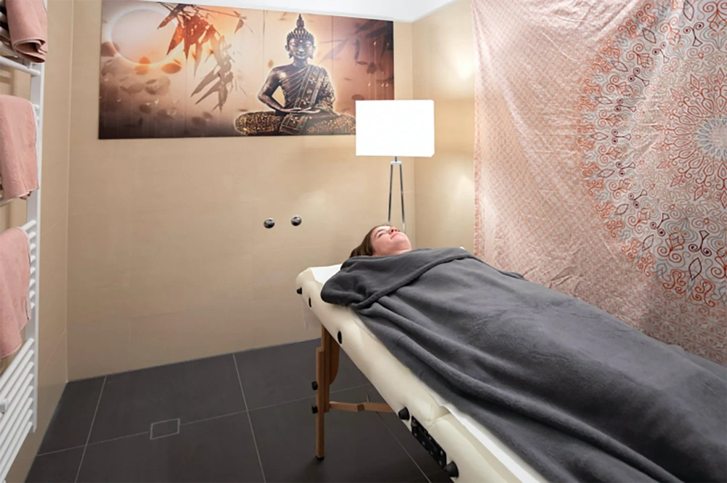 Eine Person liegt auf einer Behandlungsliege in einem Raum, der für Wellness oder Massage eingerichtet ist, mit einem Buddha-Bartempel-Poster und einer Stehlampe im Hintergrund.