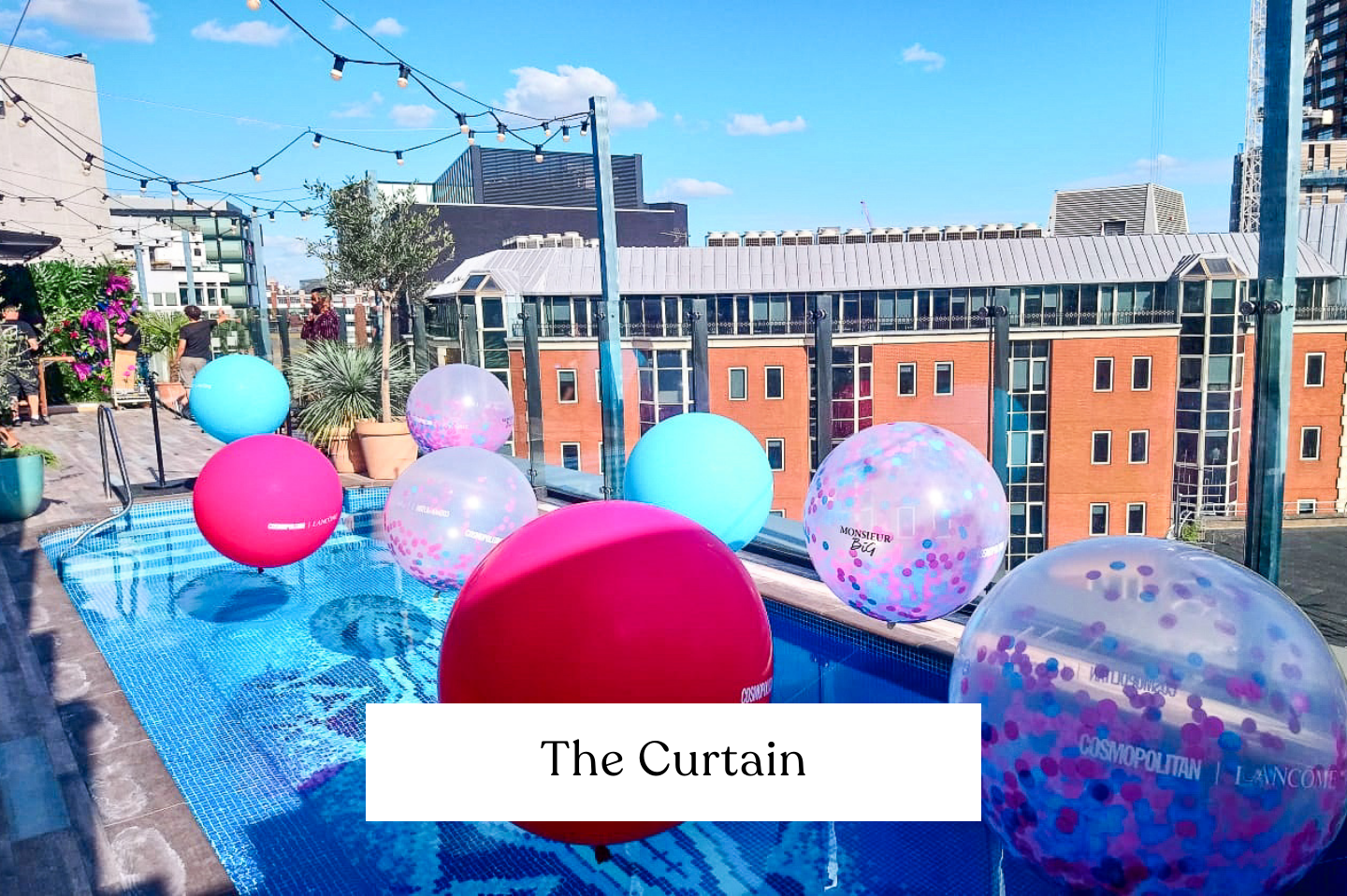 On a rooftop pool with large pink, blue, and confetti giant balloons floating on the water, city buildings and string lights visible in the background.
