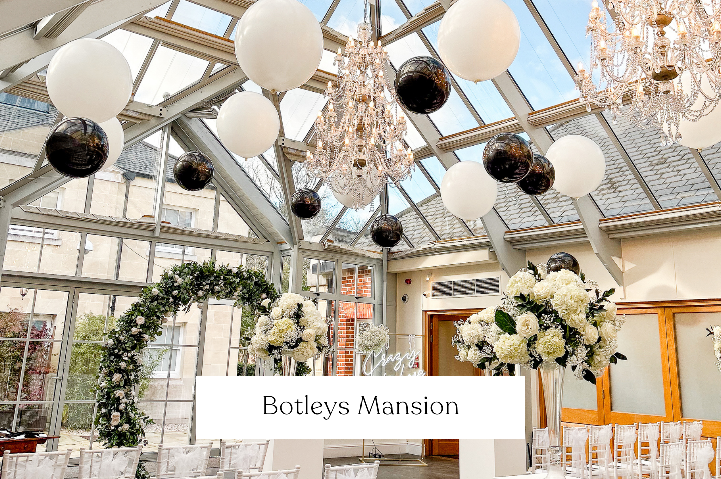 Elegant indoor wedding suspended ceiling balloon setup at Botleys Mansion with white floral centerpieces, chandeliers, black and white balloons, and decorative arches in a glass-roofed conservatory.