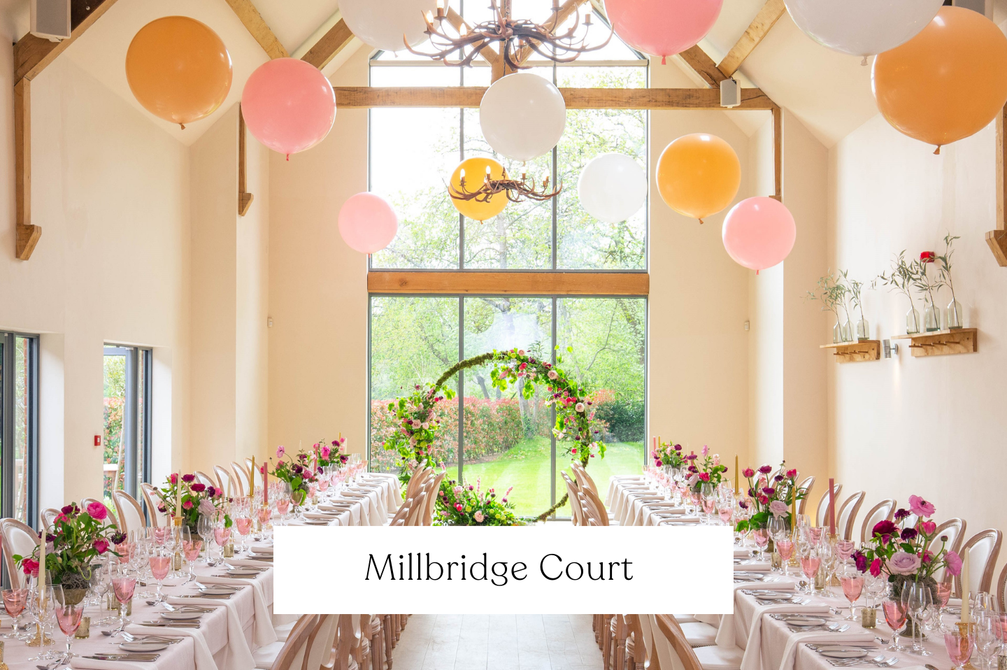 Elegant wedding reception setup at Millbridge Court, Hampshire, decorated with pink and purple floral centerpieces, arranged beneath large pink, orange, and white balloons, and a floral arch at the large window overlooking a lush garden.