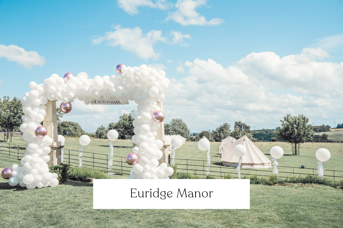 Outdoor wedding event setup with a balloon arch made of white and metallic pink balloons, a glastonbury festival theme with tents in the background.