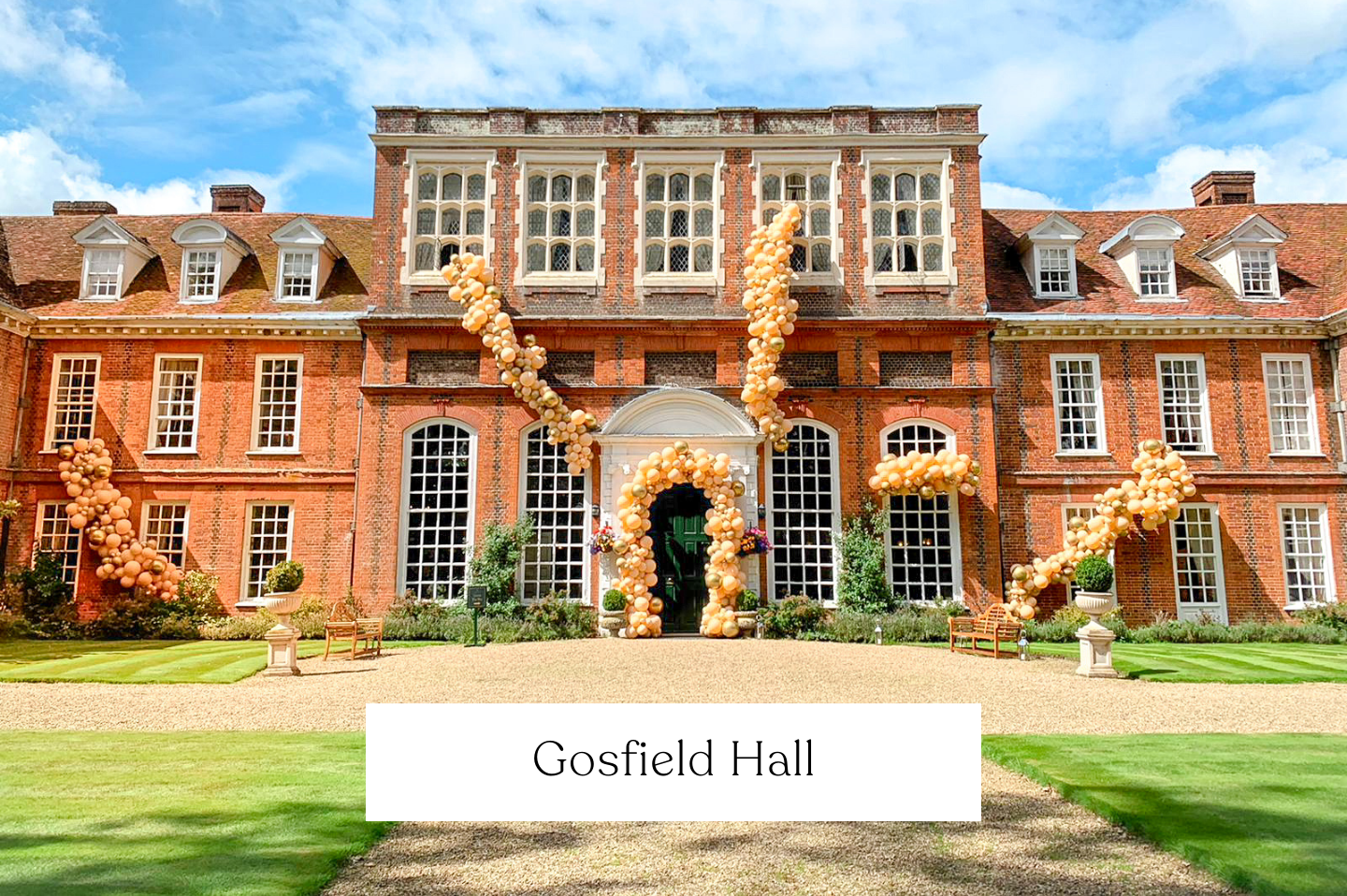 Gosfield Hall decorated with peach-coloured balloons forming a large arch over the entrance and trailing along the sides. Asymmetrical Balloon Arch.