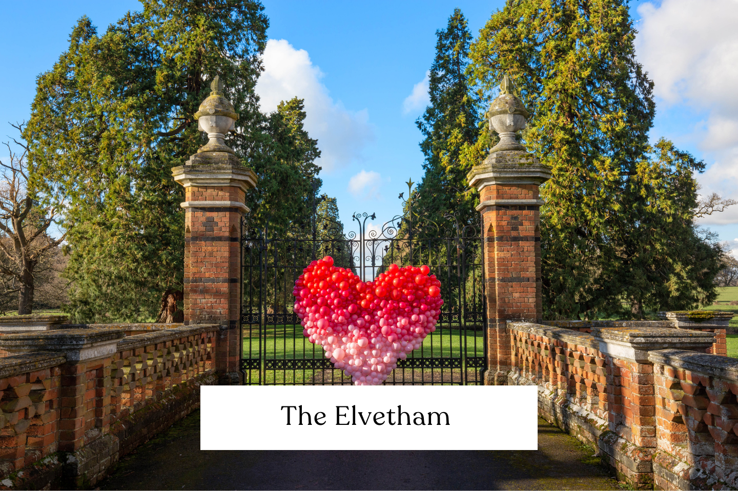 A heart-shaped balloon display with red, pink at The Elvetham estate, framed by brick pillars and lush green trees under a blue sky. Perfect for Valentine's day large scale unique gift. 