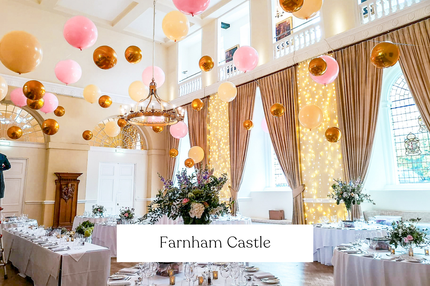 Decorated banquet wedding hall at Farnham Castle with pink, gold, and peach balloons, string lights, flower arrangements, and set tables for an event.