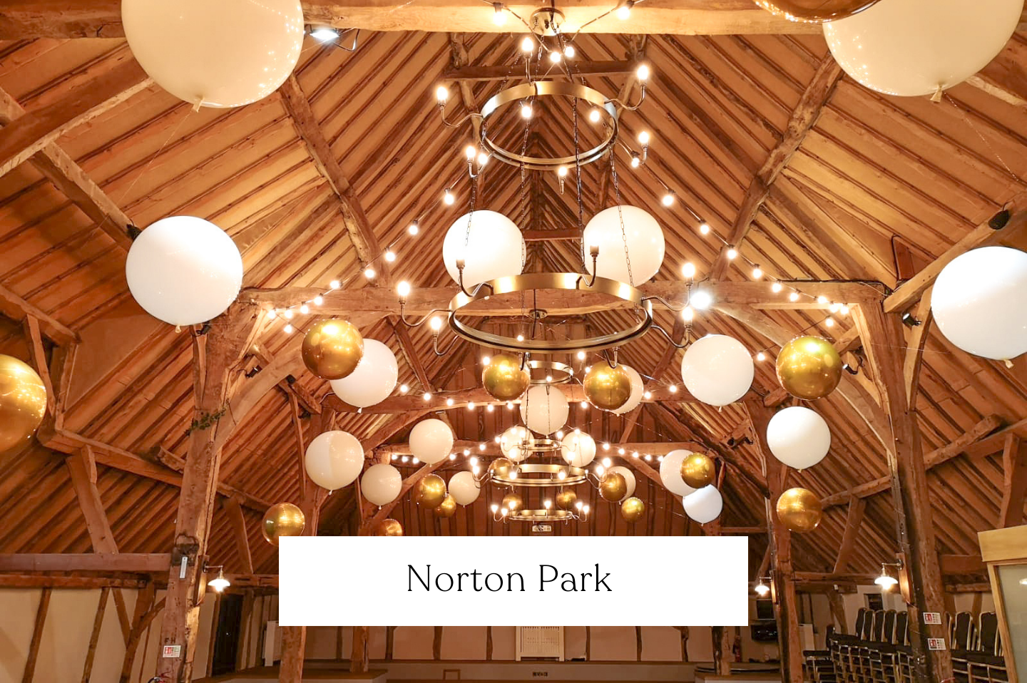 Decorative ceiling with hanging white and gold spherical balloons, warm string lights, and chandeliers inside a rustic wooden barn with exposed beams.