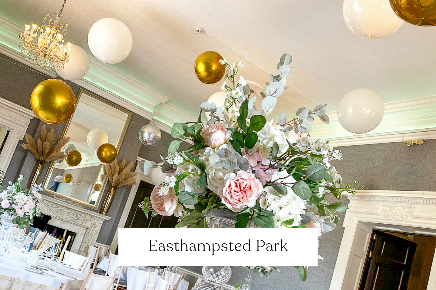 Elegant event room decorated with white and gold balloons, large floral centerpiece with pink and white roses and greenery.