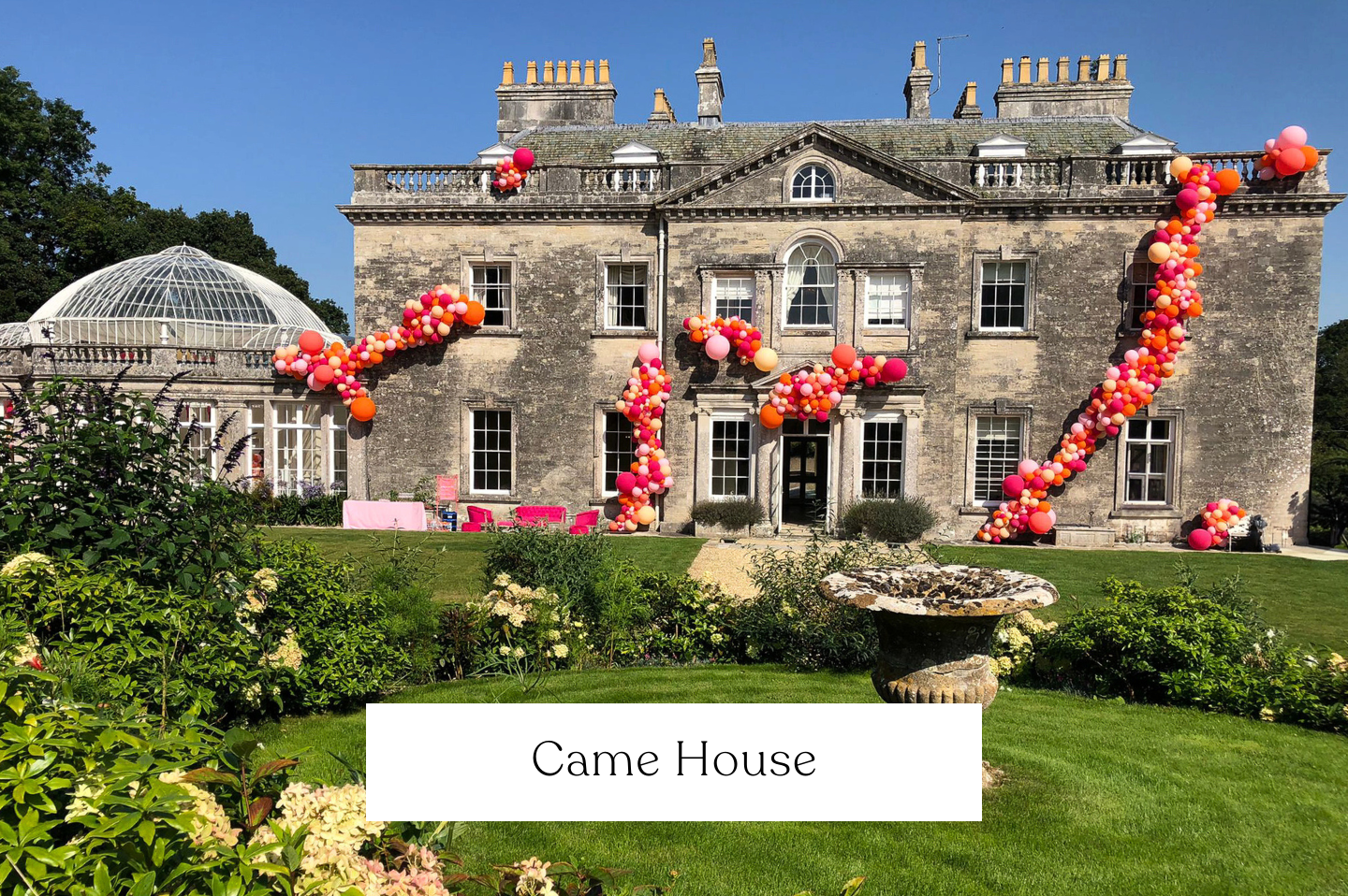A large historic stone mansion decorated with external garlands in pink, red, and orange balloons for a celebration.