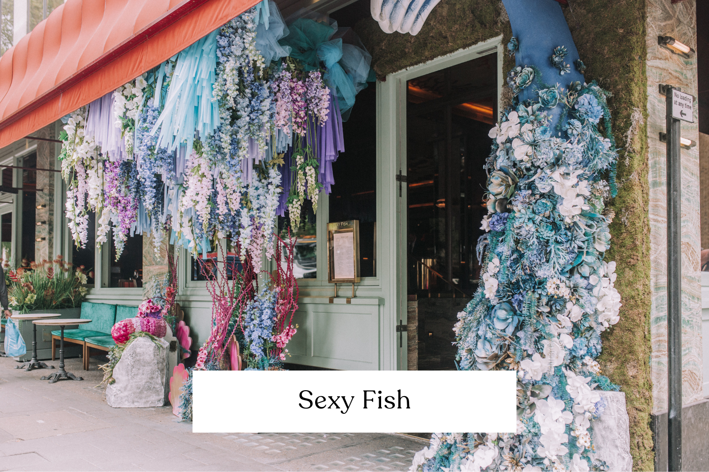 The entrance of a restaurant called Sexy Fish decorated with an elaborate display of blue and white flowers and balloons within the floral arrangement. 
