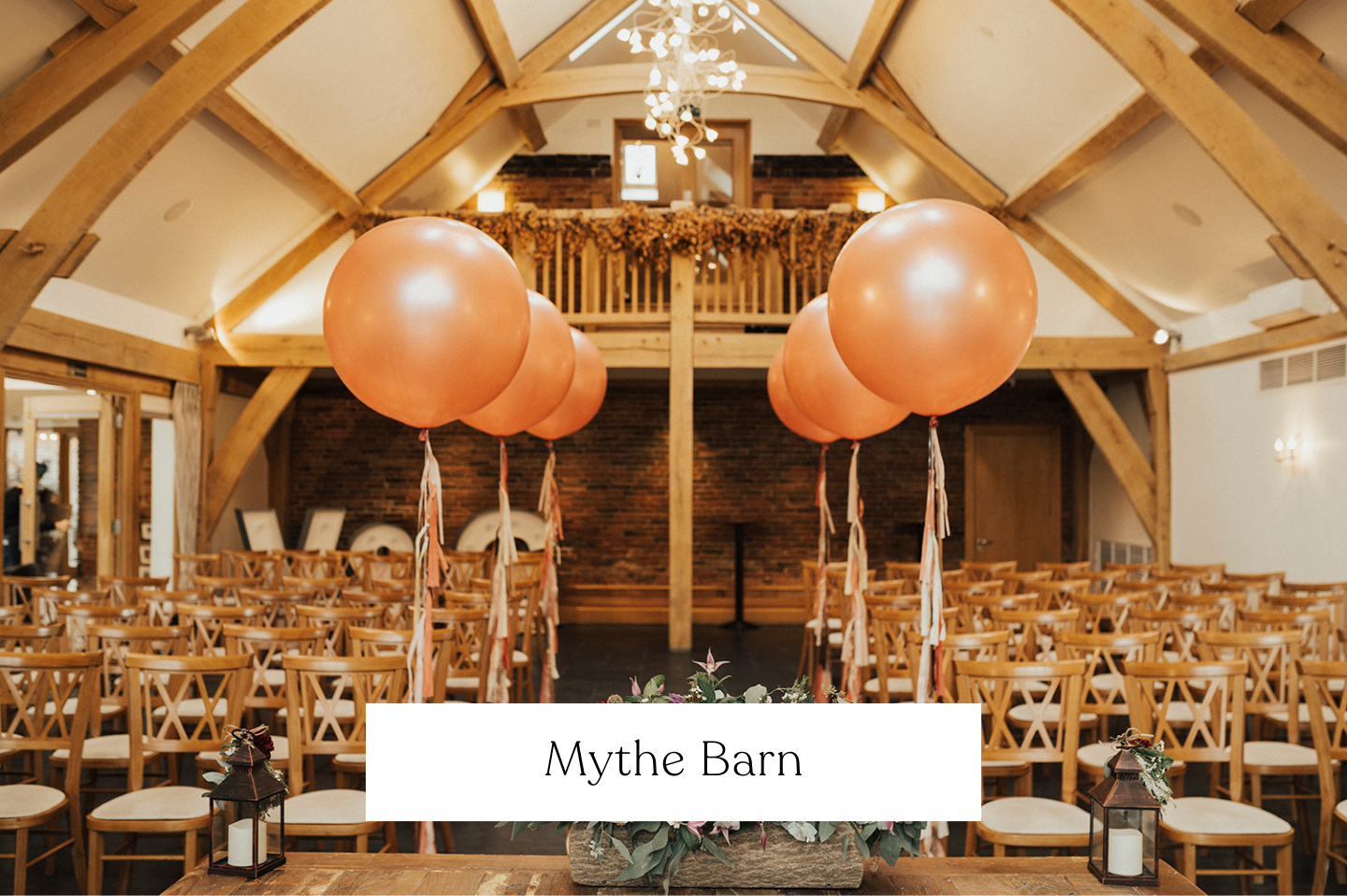 Wedding ceremony setup inside Mythe Barn with large pink balloons down the aisle, wooden chairs, floral centerpiece, lanterns, and a brick wall at the front.