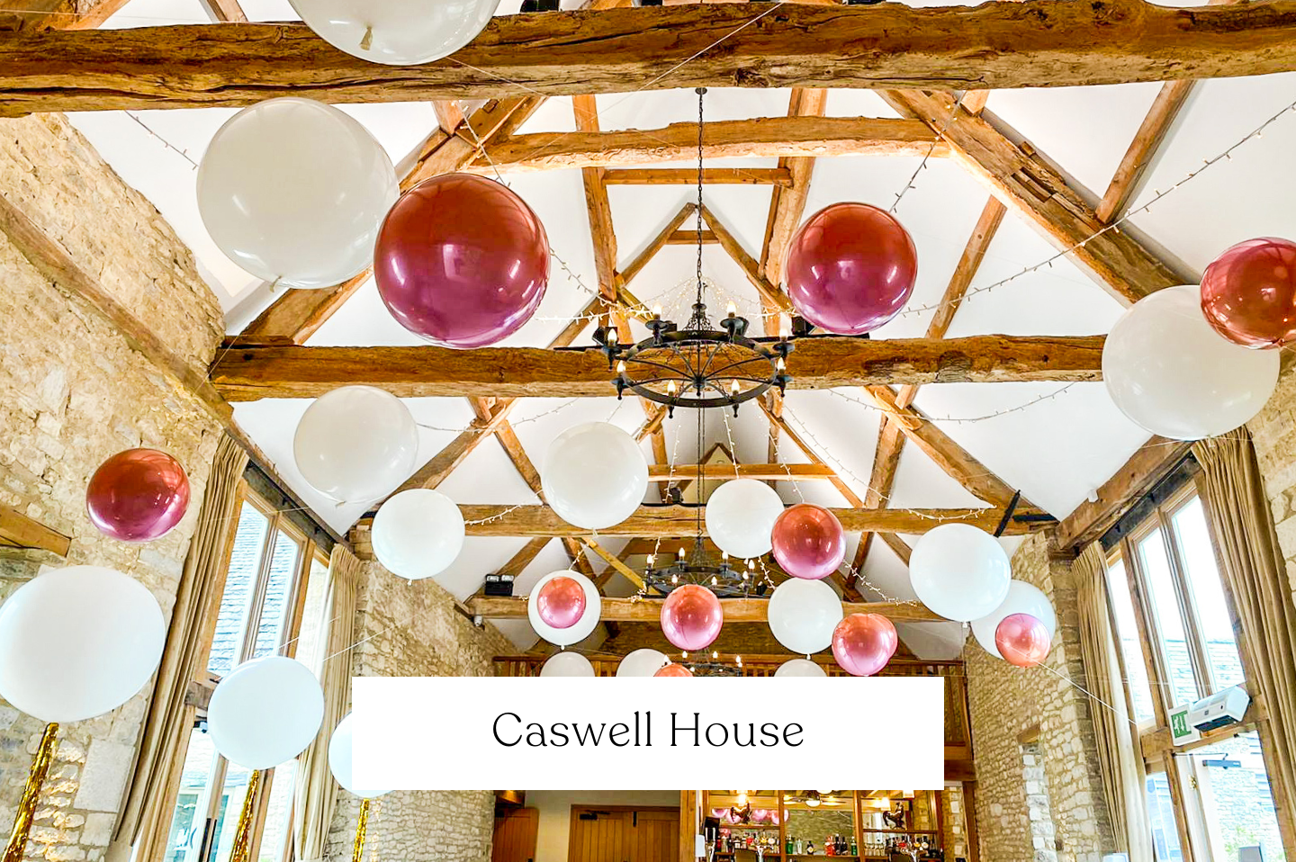 Interior of a barn wedding with exposed wooden beams, stone walls, and large windows, decorated with suspended floating white and pink balloons.