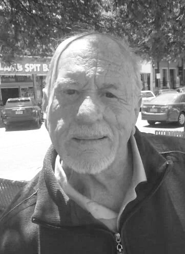Older man with short hair, beard, wearing a jacket and collared shirt, standing outdoors in a parking lot with cars and a building in the background.