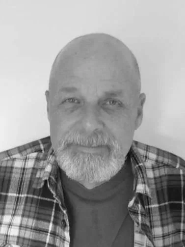 A black and white photo of a middle-aged man with a beard, wearing a plaid shirt and a t-shirt underneath, looking directly at the camera.