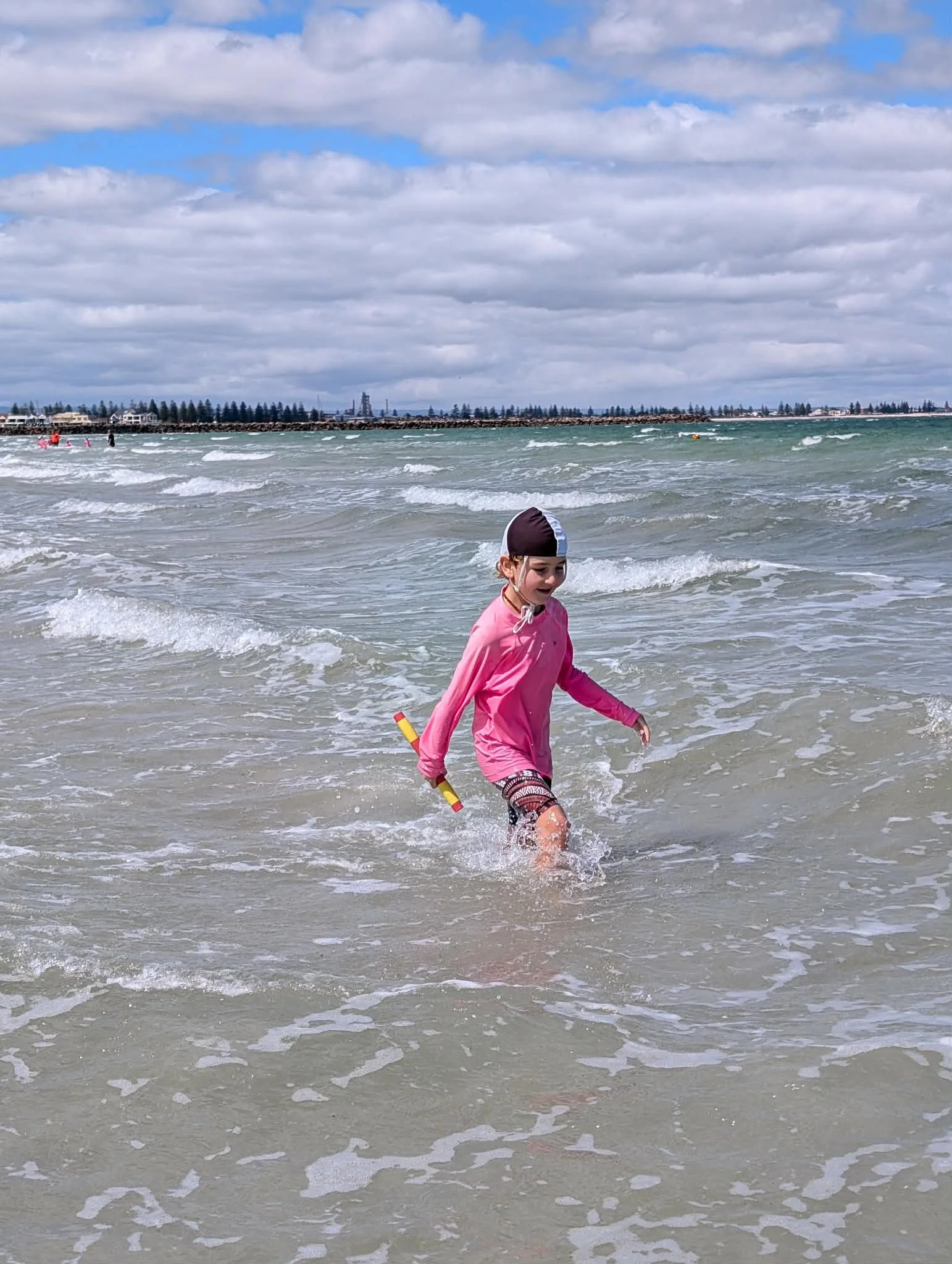 A young girl in a pink long-sleeve shirt, patterned shorts, and a black and white swim cap playing in the shallow water at the beach, holding a toy above her shoulder with waves crashing around her.