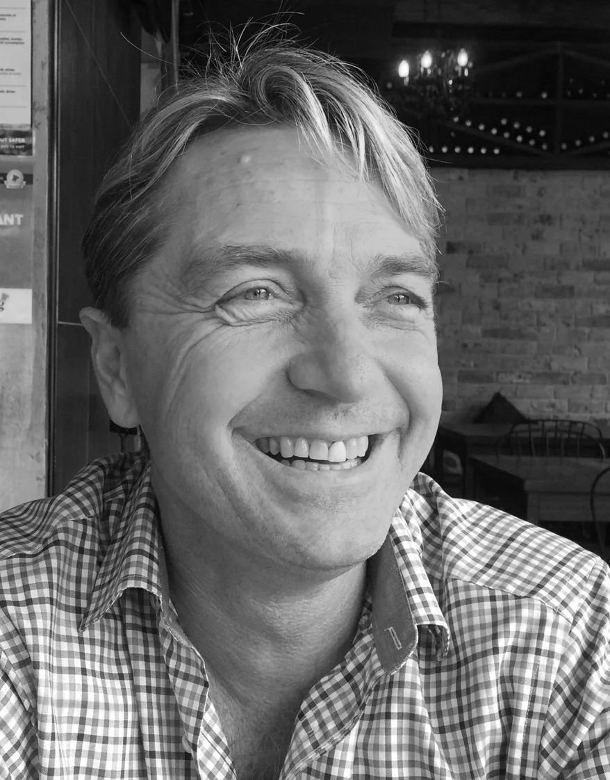 A smiling man with light-colored hair and a checkered shirt sitting in a restaurant or cafe with wooden walls and a chandelier visible in the background.