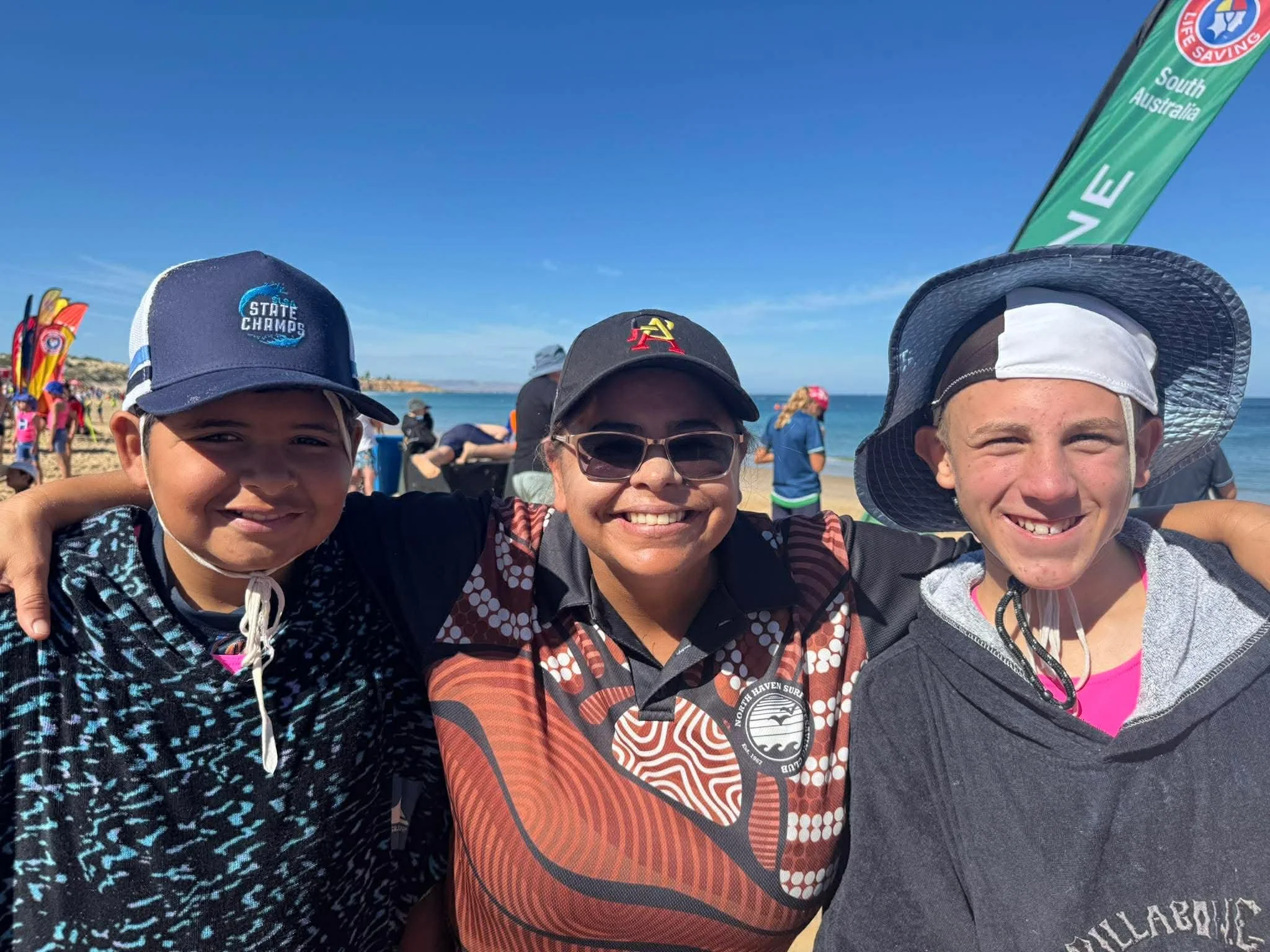 Three smiling people, two children and one woman, with arms around each other, standing on a beach with blue sky, ocean, and flags in the background.