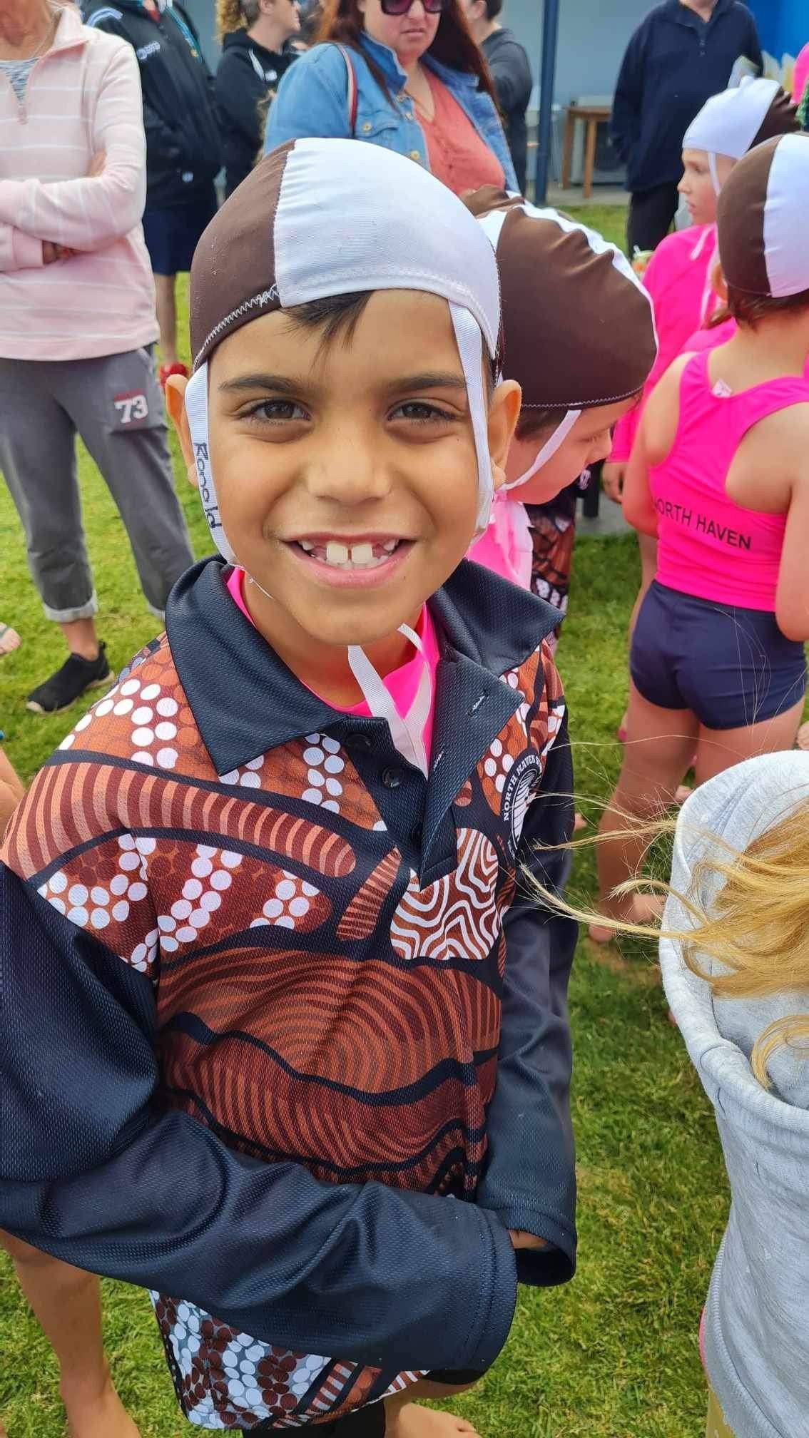 Smiling young boy with missing front teeth, wearing a helmet and colorful sports jacket, standing outdoors with other children and adults in the background.