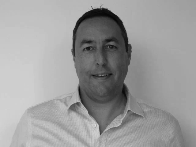 A black and white photo of a man with short dark hair, wearing a collared shirt, smiling slightly, standing against a plain light background.
