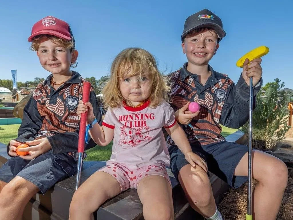 Three children sitting outdoors on a sunny day, holding golf clubs and golf balls, with a playground in the background.