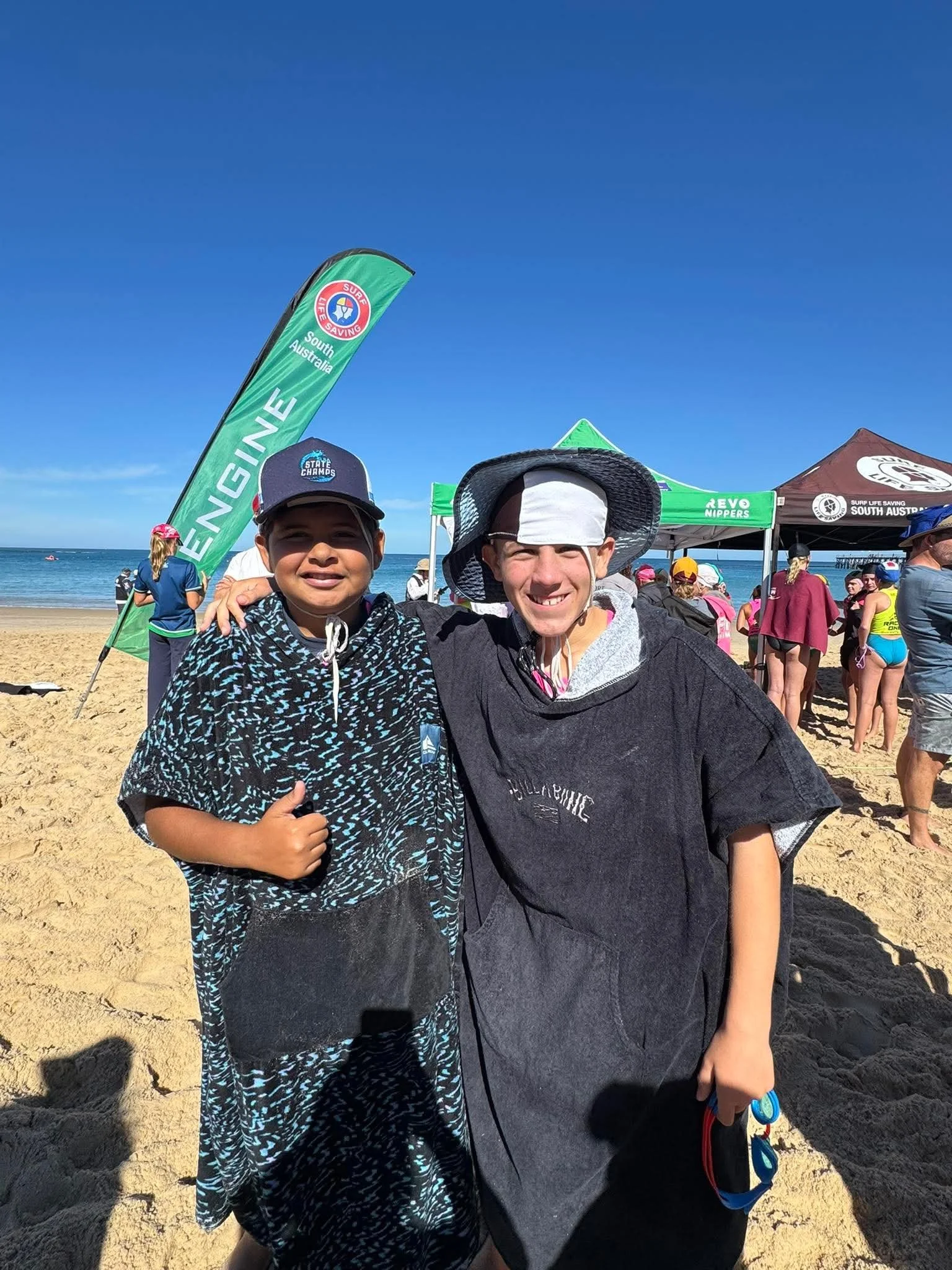 Two boys standing on a sandy beach, smiling and posing for the camera, with a group of people and event tents in the background, under a clear blue sky.