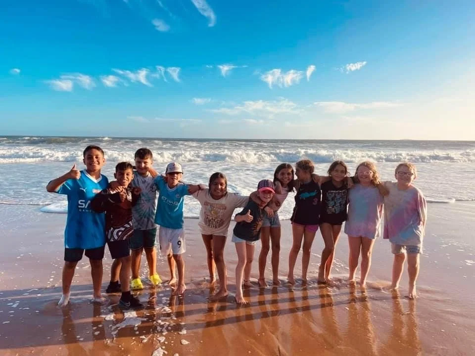 A group of eleven children standing together at the beach with waves in the background, smiling and enjoying the sunny day.