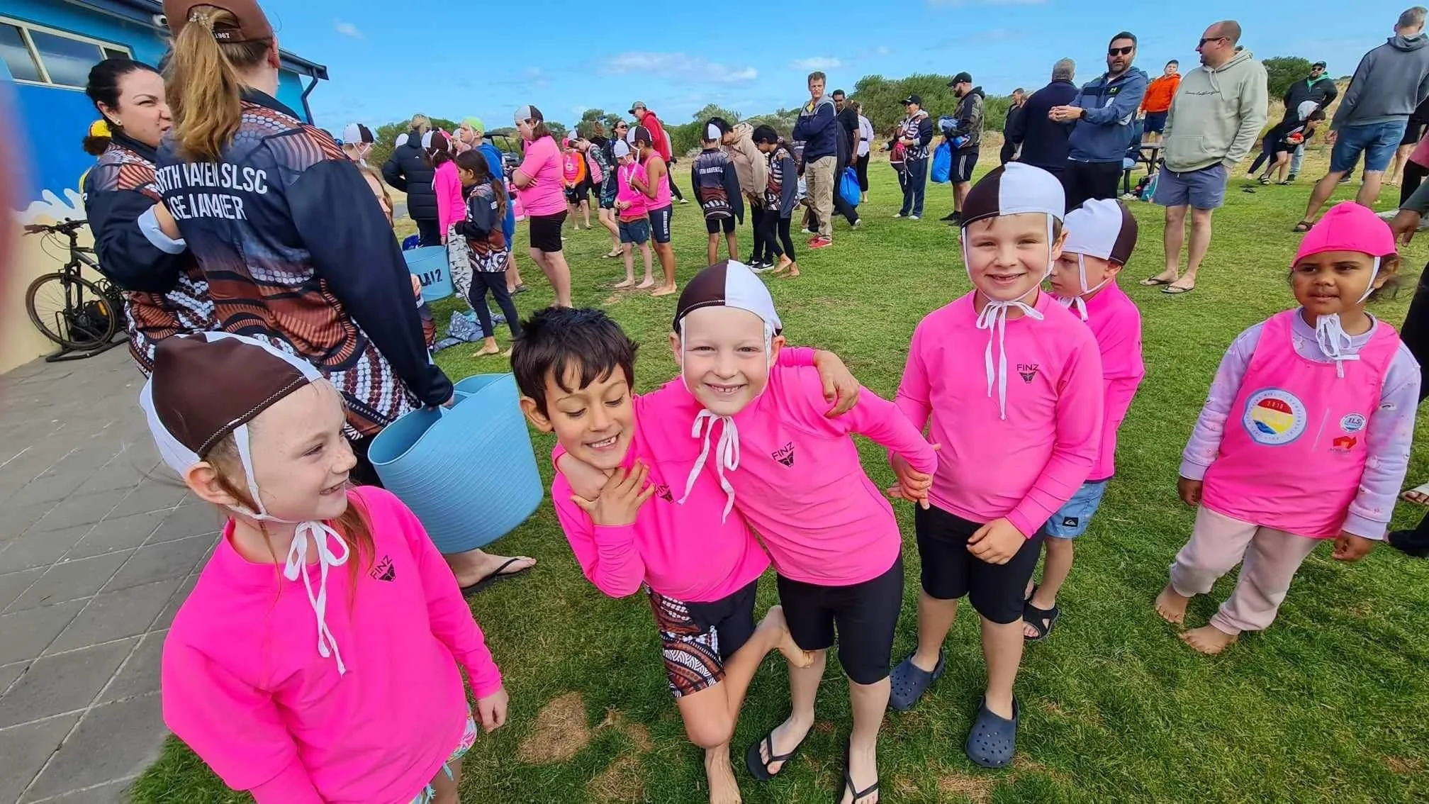Group of children in pink swimwear and hats, smiling and embracing, at an outdoor event with many people and tents in the background.