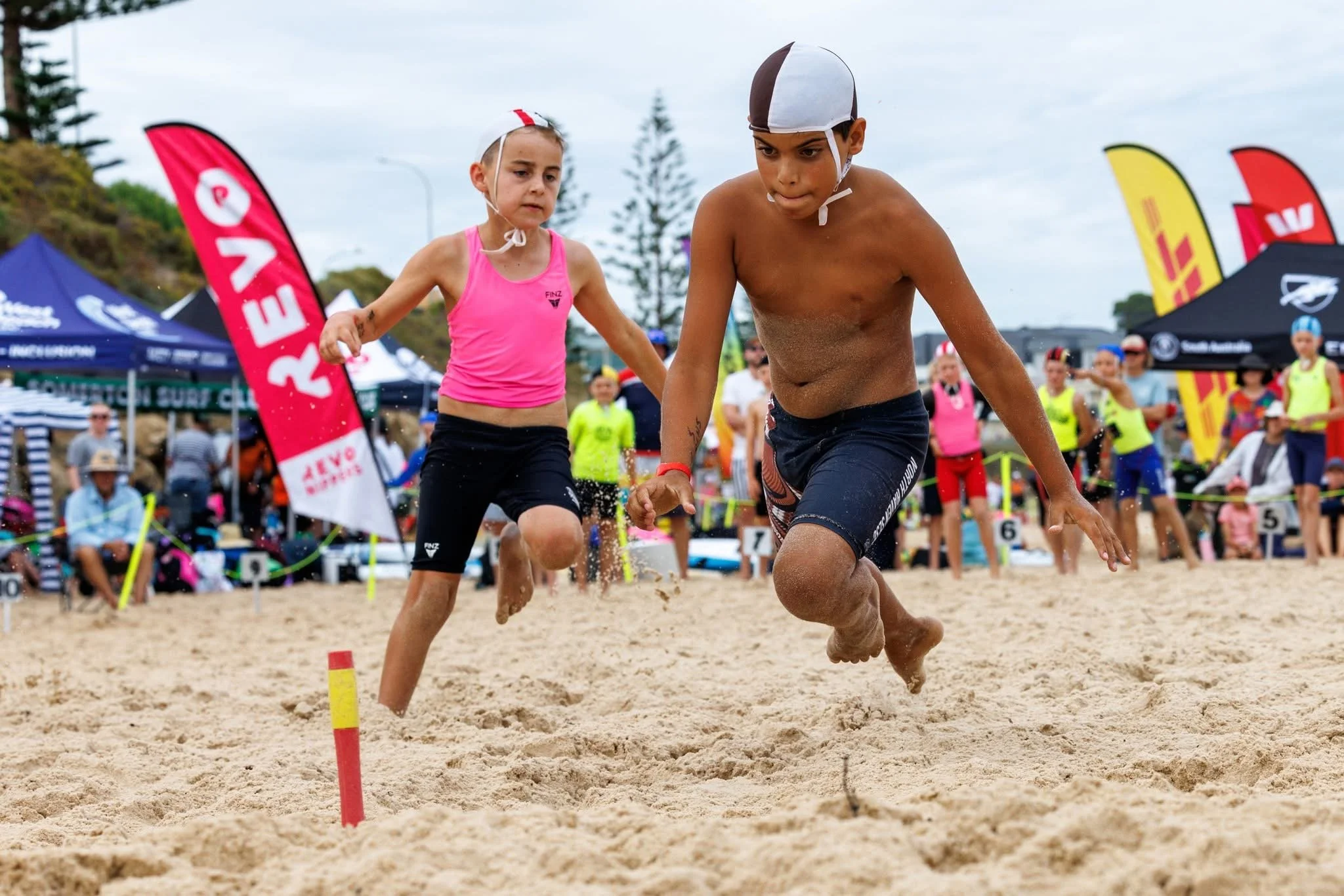 Two young kids running on a sandy beach during a sporting event with a crowd and tents in the background.