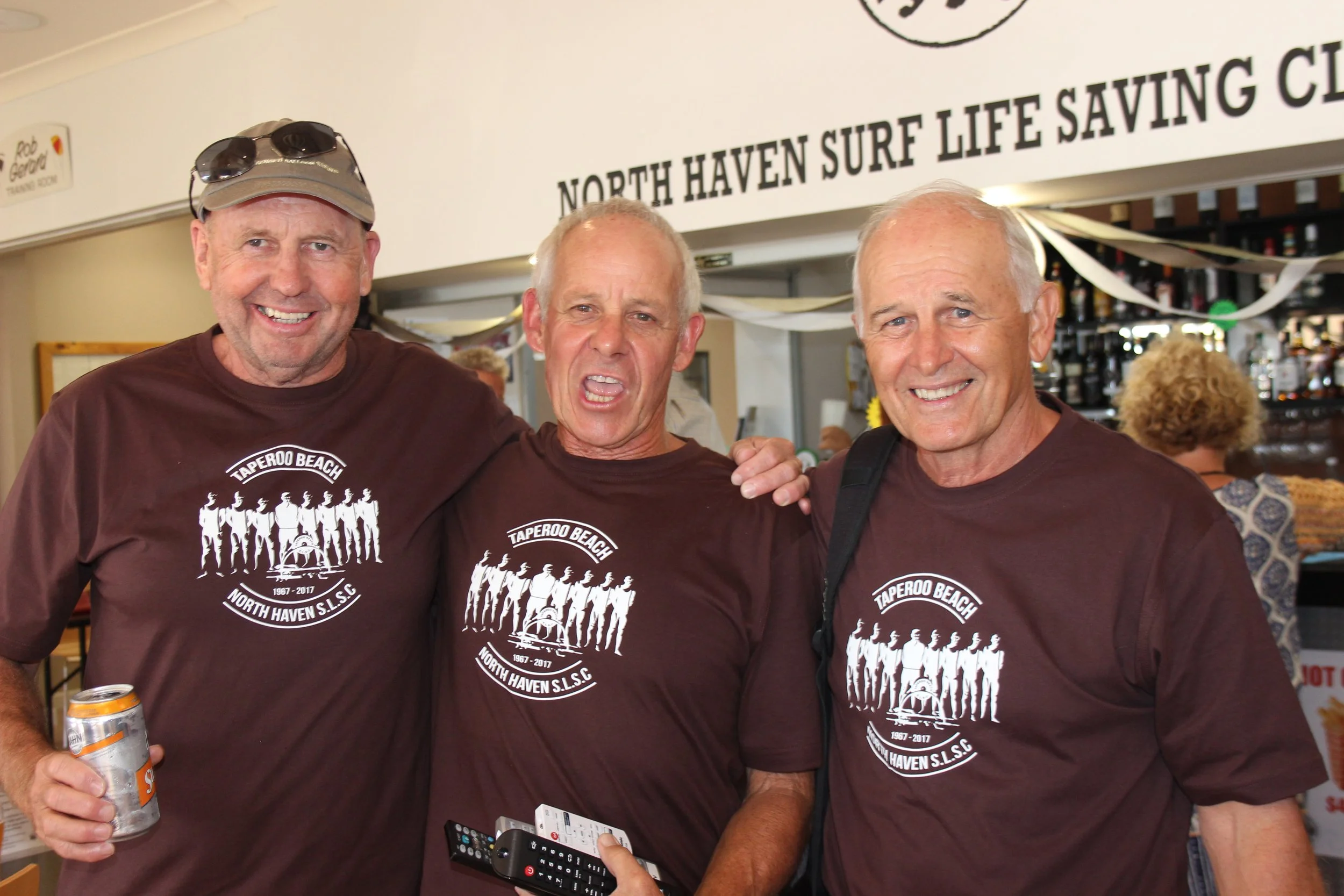 Three smiling men in matching brown T-shirts with a logo that says 'Taperoo Beach North Haven S.L.S.C.' standing indoors, with one holding a soda can and another holding a remote control, at a bar or social event.