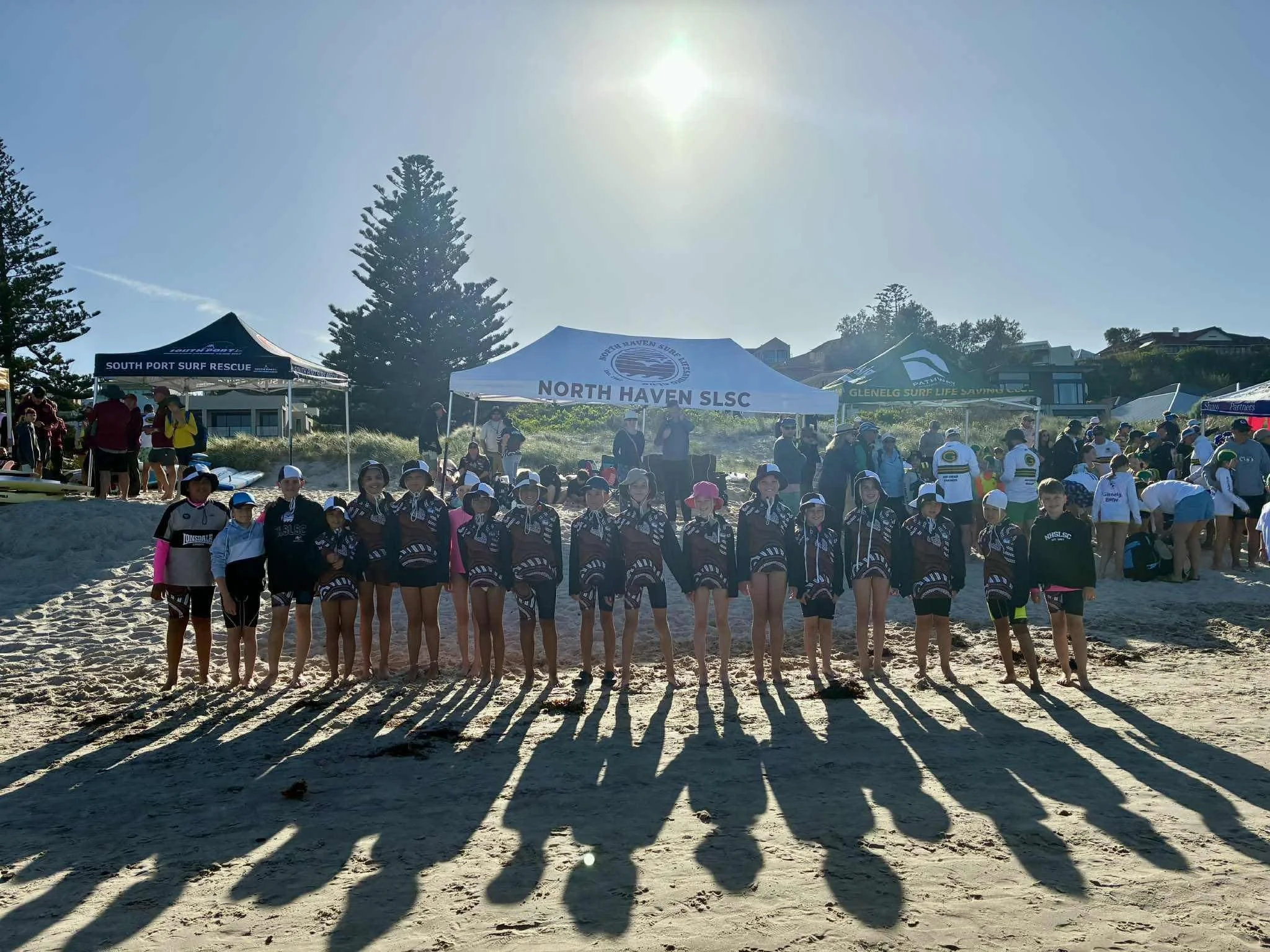 Group of young female surf lifesavers in uniform, standing on a beach with long shadows, at a surf rescue event with tents in the background.
