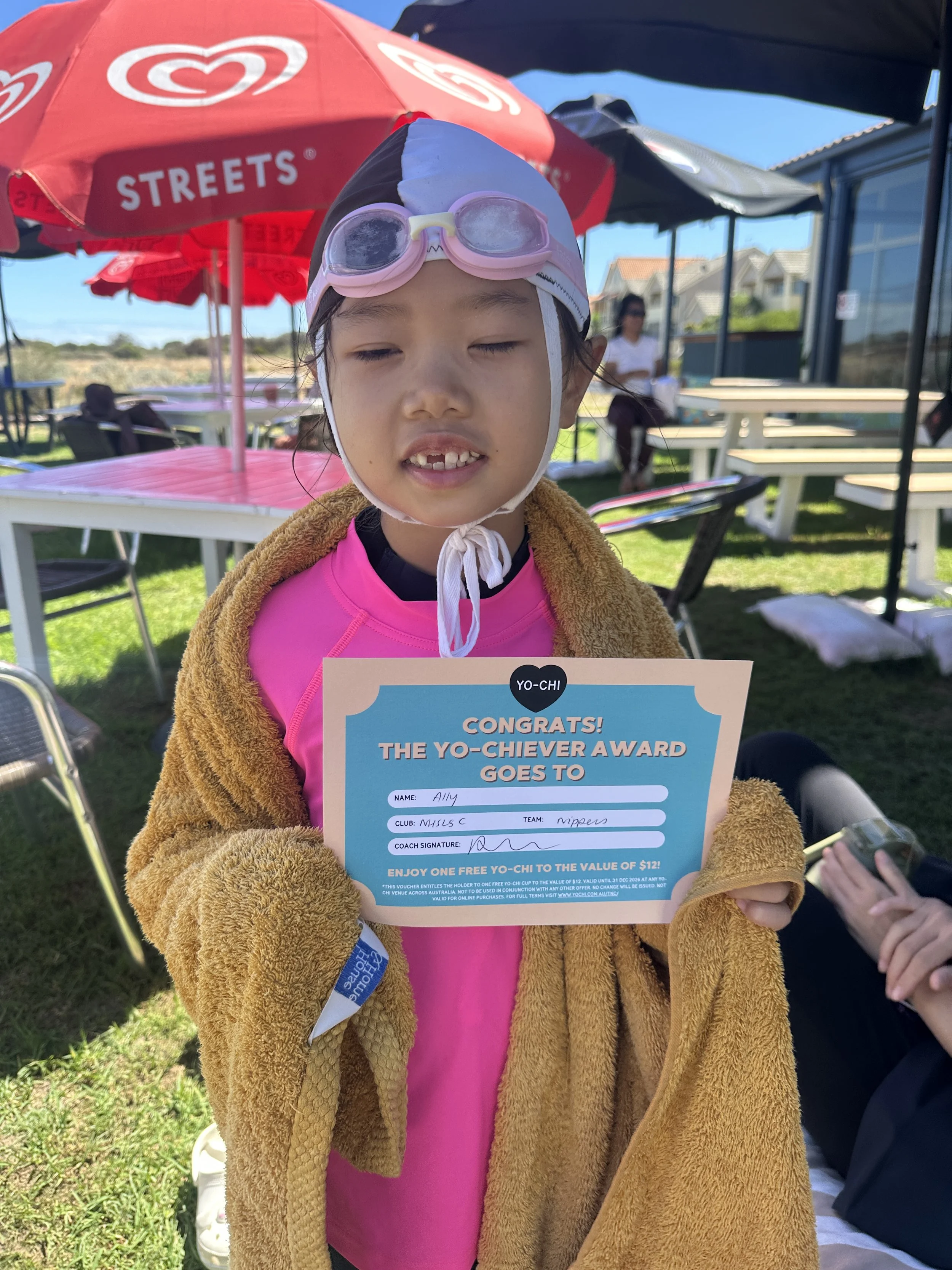 Young girl in swimming gear holding a certificate outdoors at a sunny location with tables and umbrellas in the background.