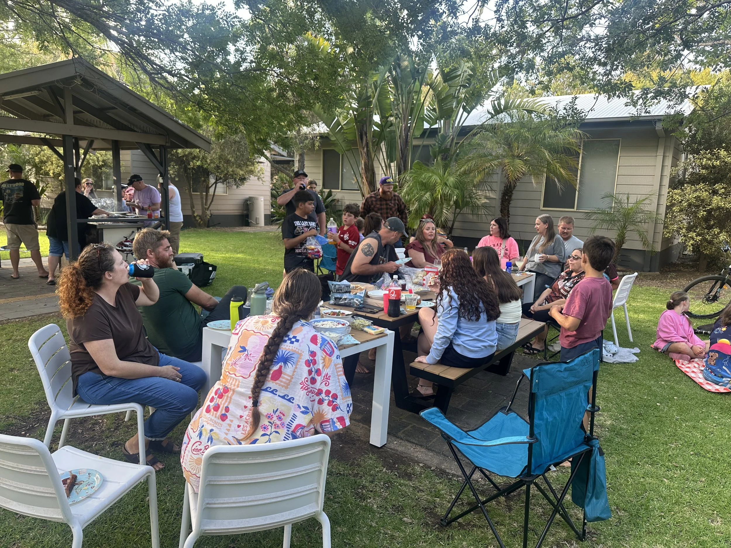 A group of people gathered outdoors in a backyard, sitting and standing around tables with food and drinks, enjoying a social gathering or celebration on a sunny day.